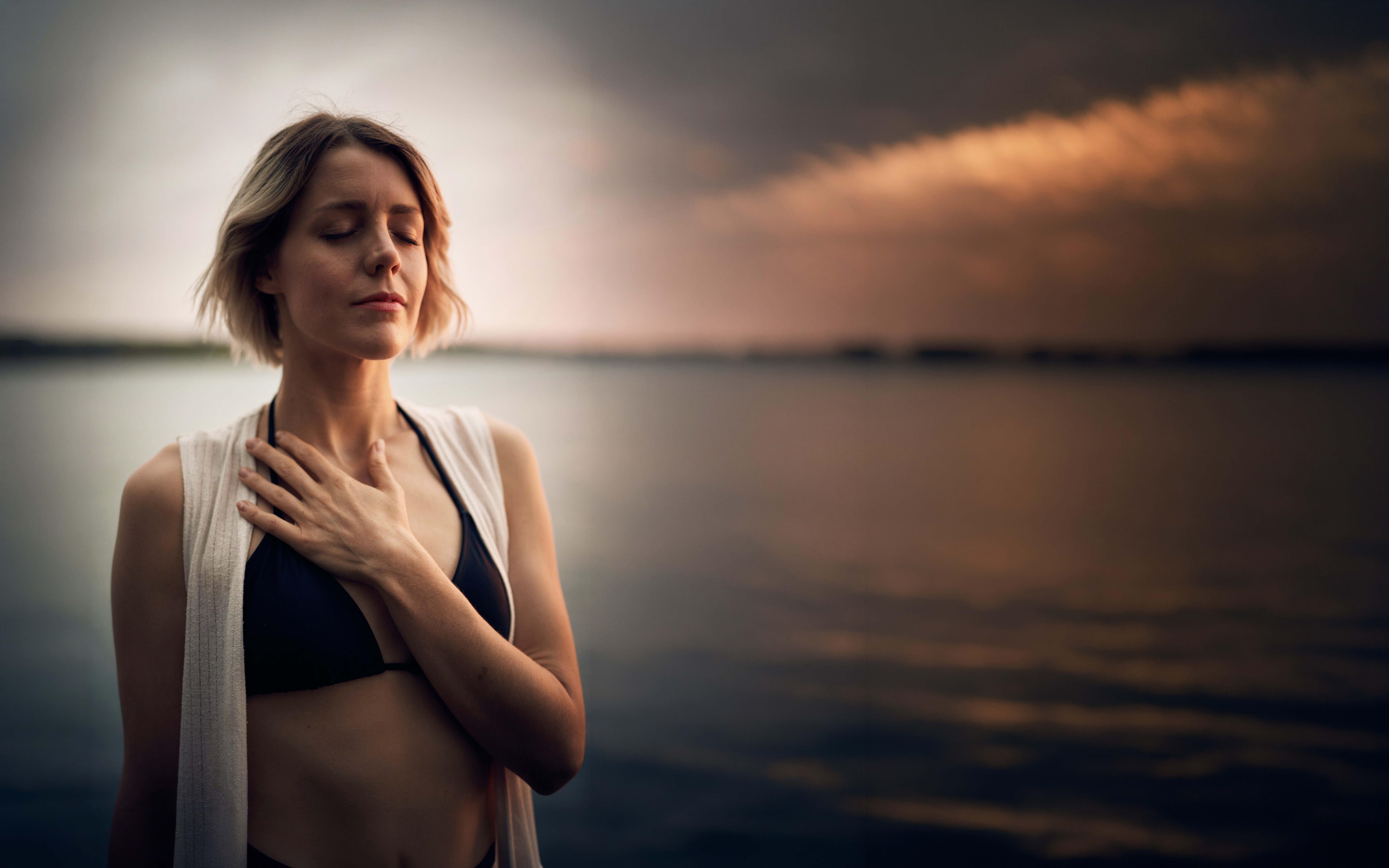 Woman practising mindful breathing beside open water at sunset, with a tranquil natural backdrop.
