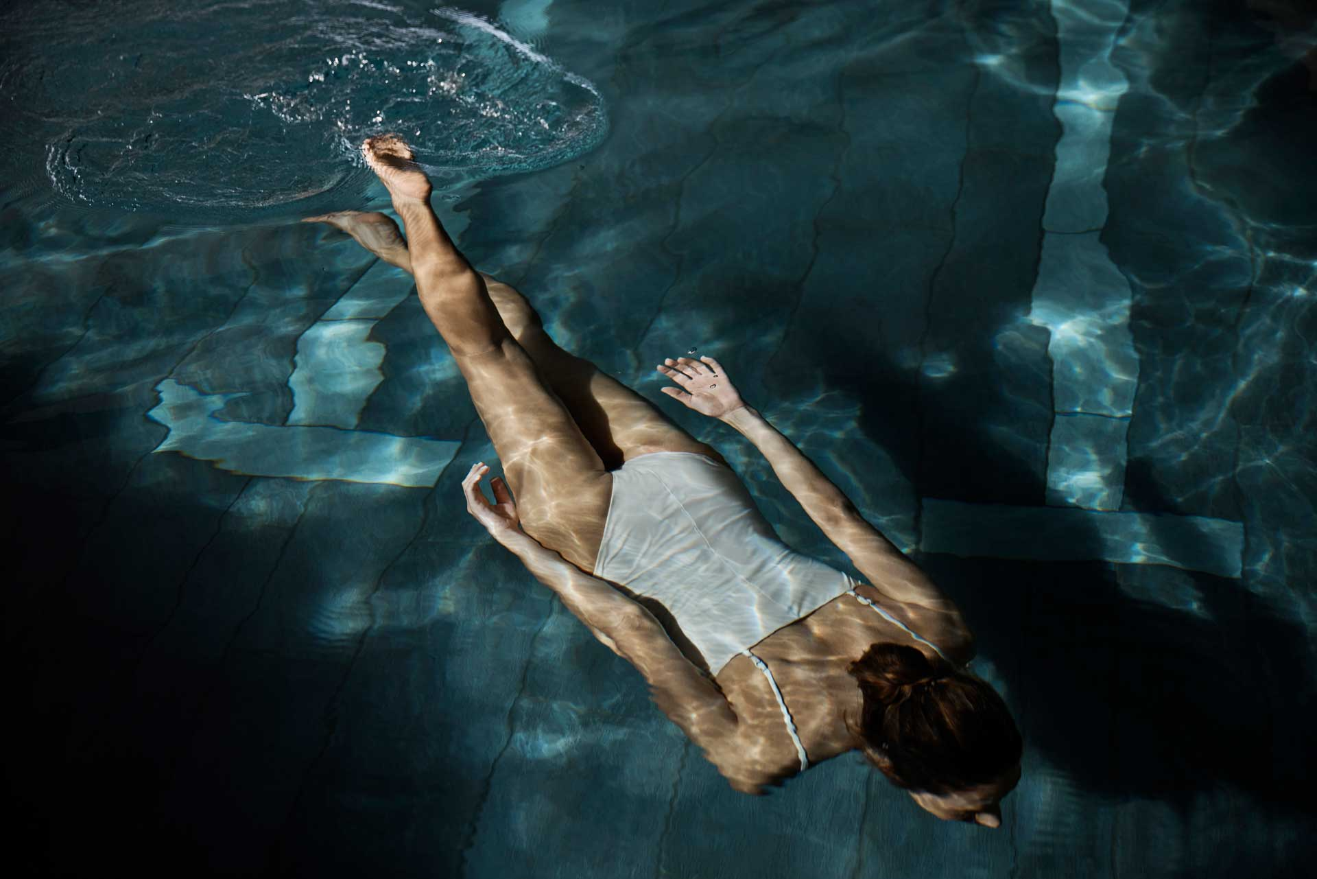 Women swimming face down underwater in a pool wearing a white swimsuit.