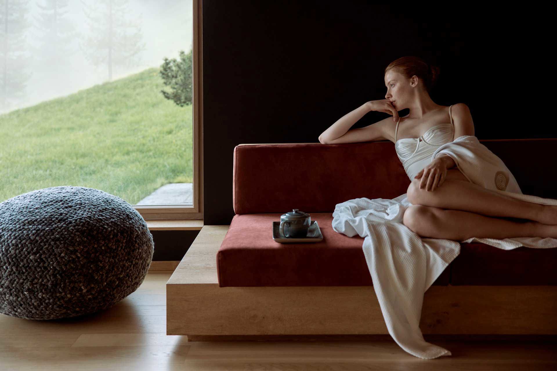 Woman relaxing on a sofa in a serene spa lounge at COMO Alpina Dolomites, gazing out towards a soft green landscape through a large window.