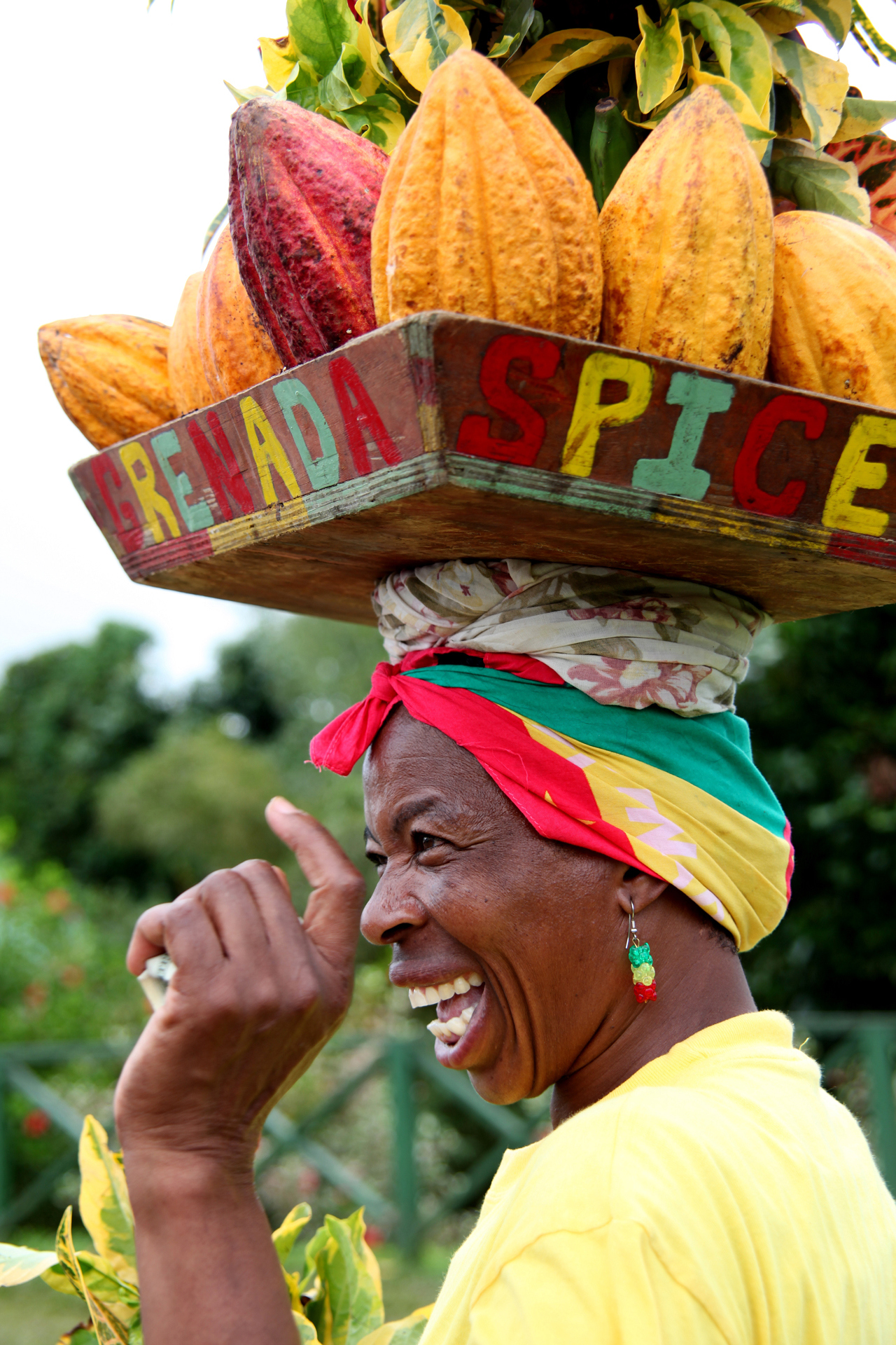A woman carrying a plate on her head which has the words Grenada Spice on it, filled with Cacao pods