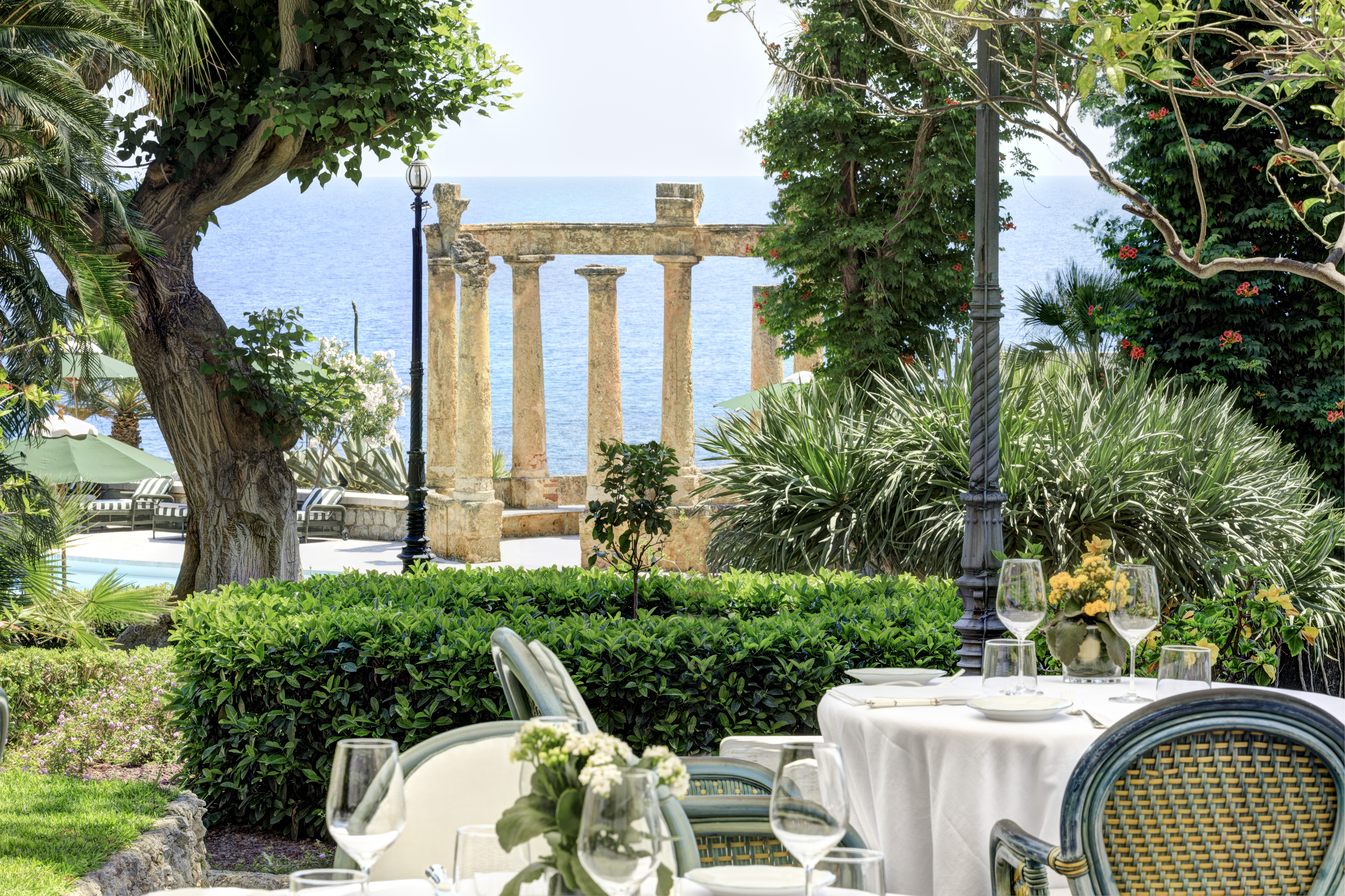 The outdoor tables of a restaurant at Villa Igiea set up for dinner with ancient ruins and the sea in the background surrounded by established greenery
