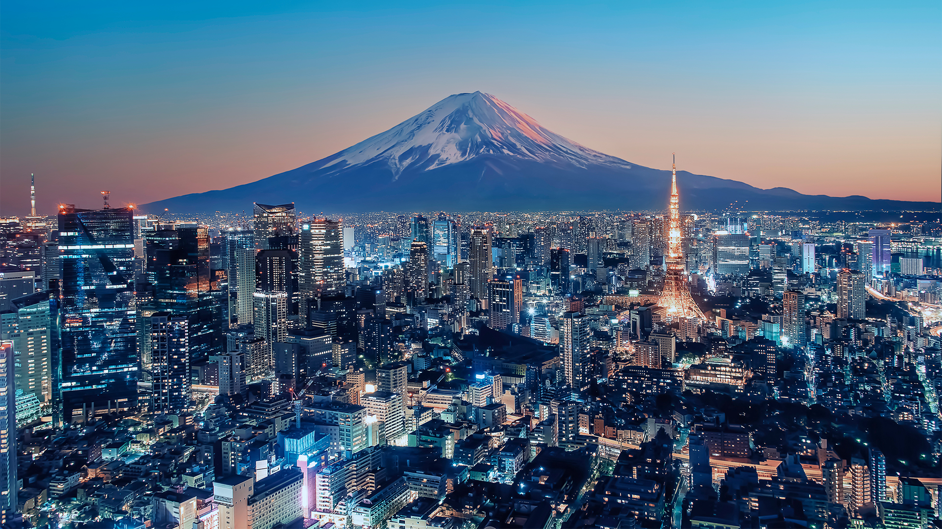 A panoramic view of a cityscape at twilight with illuminated skyscrapers, Mount Fuji in the background, and a prominent tower in the midground.