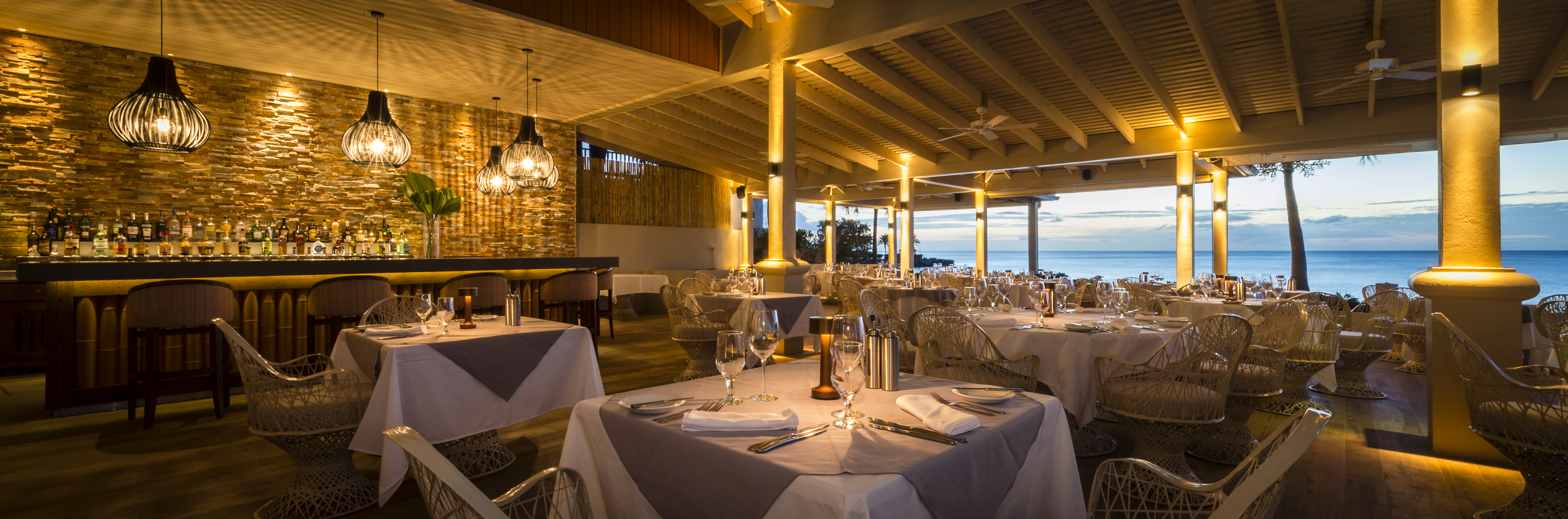 The Cove restaurant at Blue Waters in Antigua lit up at night and set up for dinner with a view of the ocean in the background