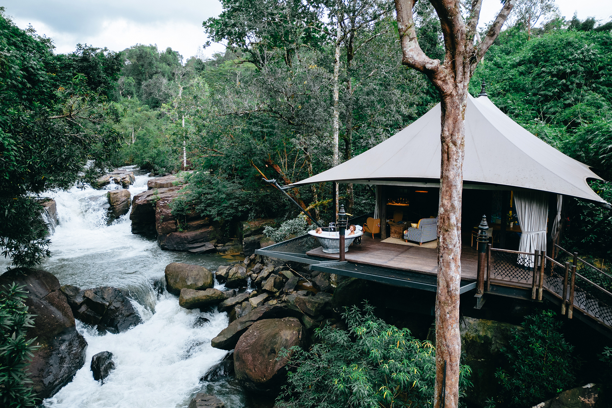 Woman in a bathtub on the terrace of a luxury tent set on a platform at Shina Mani Wild, surrounded by greenery next to a river