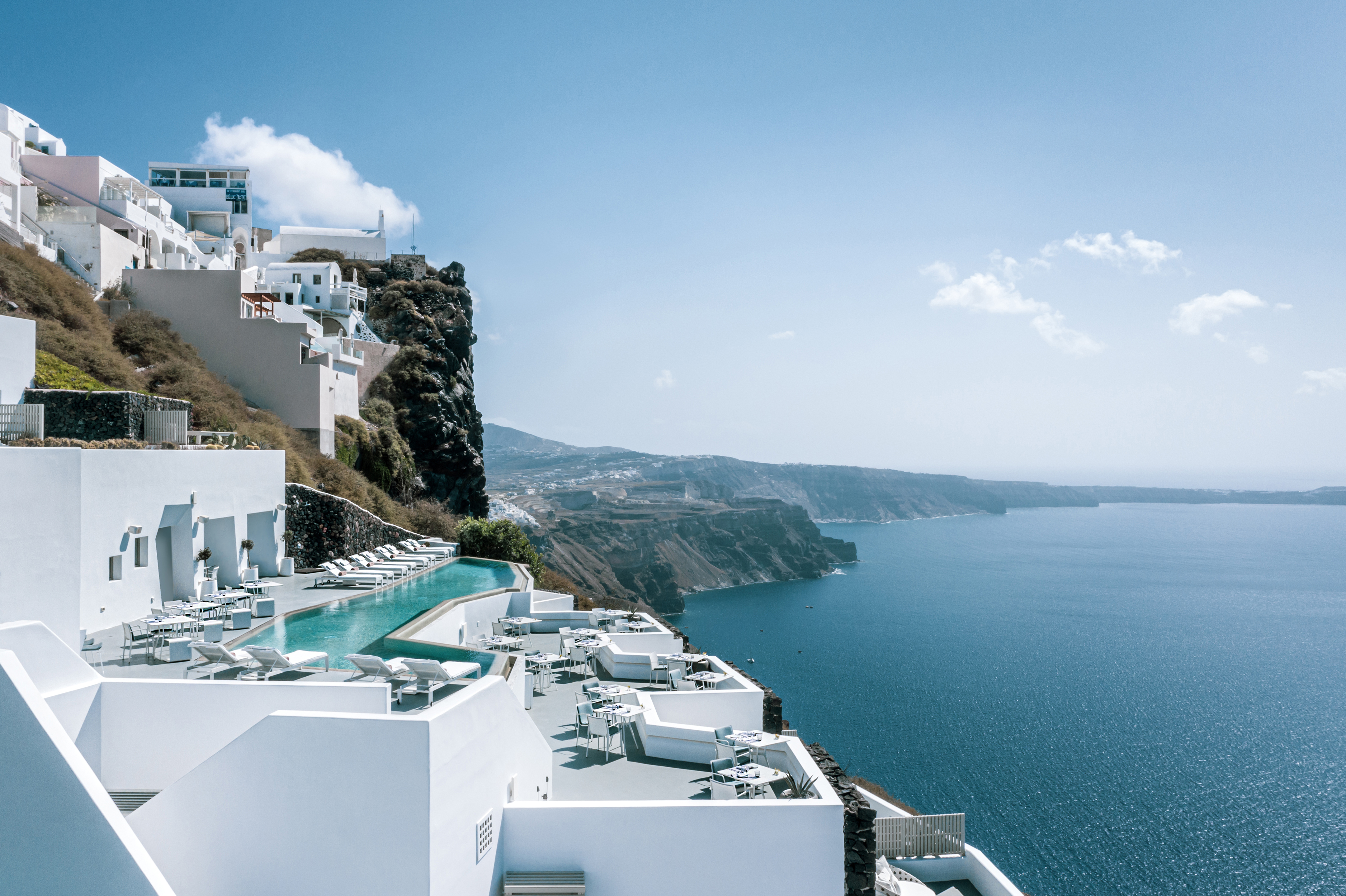 The white cliffside resort of Grace Hotel Santorini overlooking a blue ocean with a pool and lounge chairs, set against a clear sky with distant hills.