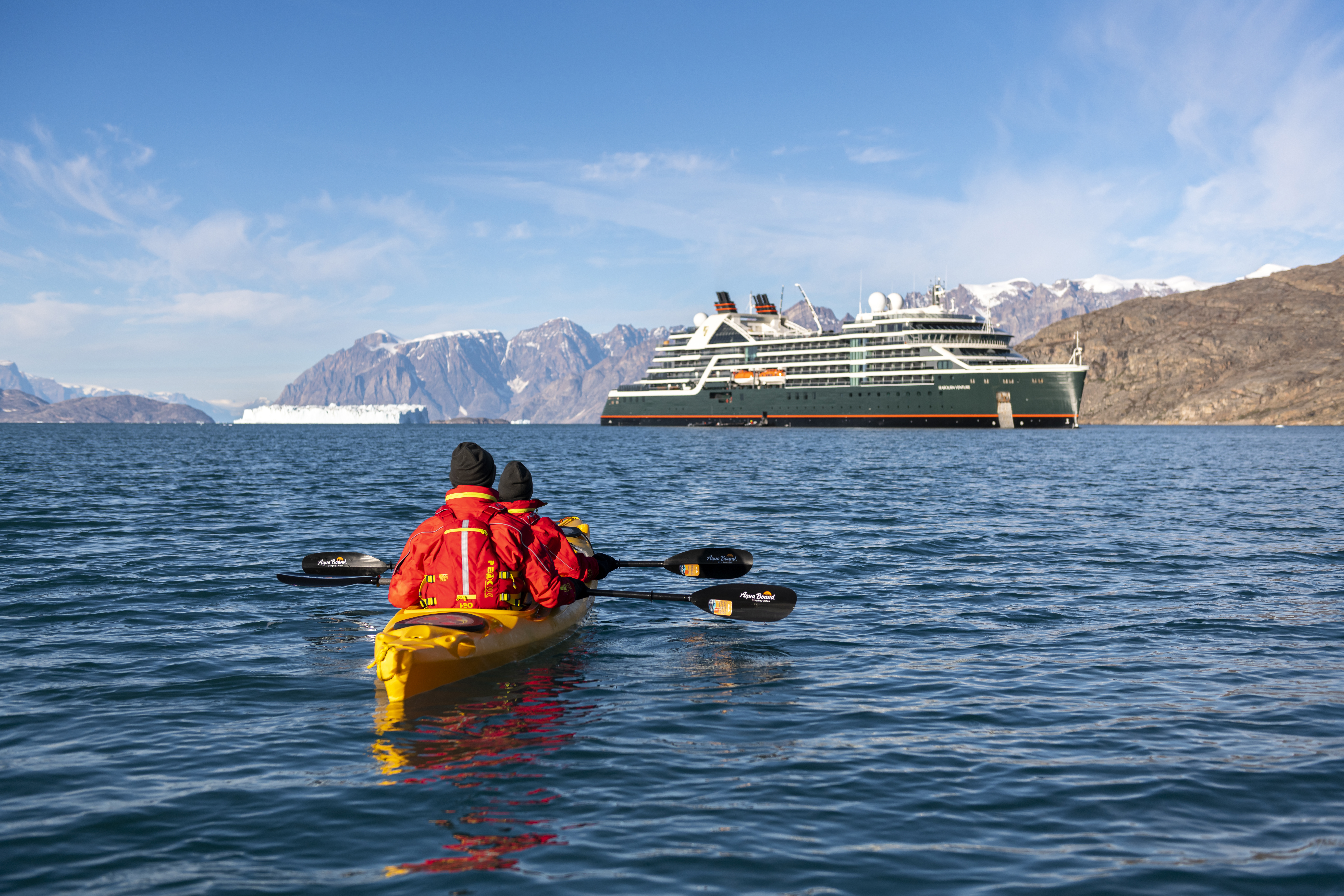 Two people in a yellow kayak paddling on calm waters towards a luxury cruise ship backed by snowy mountains