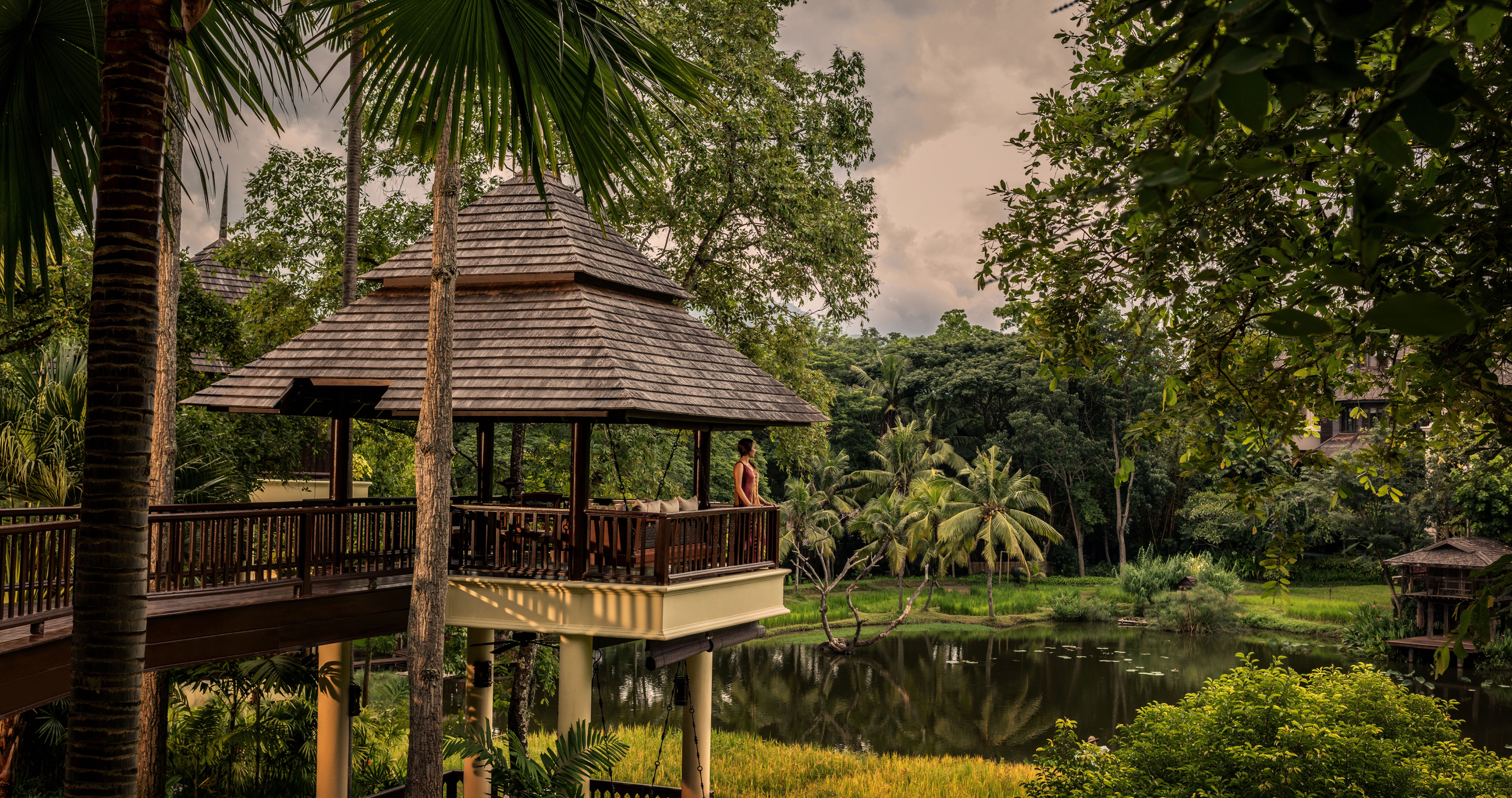 A woman standing at the end of a stilted pavillion overlooking a lake surrounded by lush greenery at Four Seasons Resort Chiang Mai
