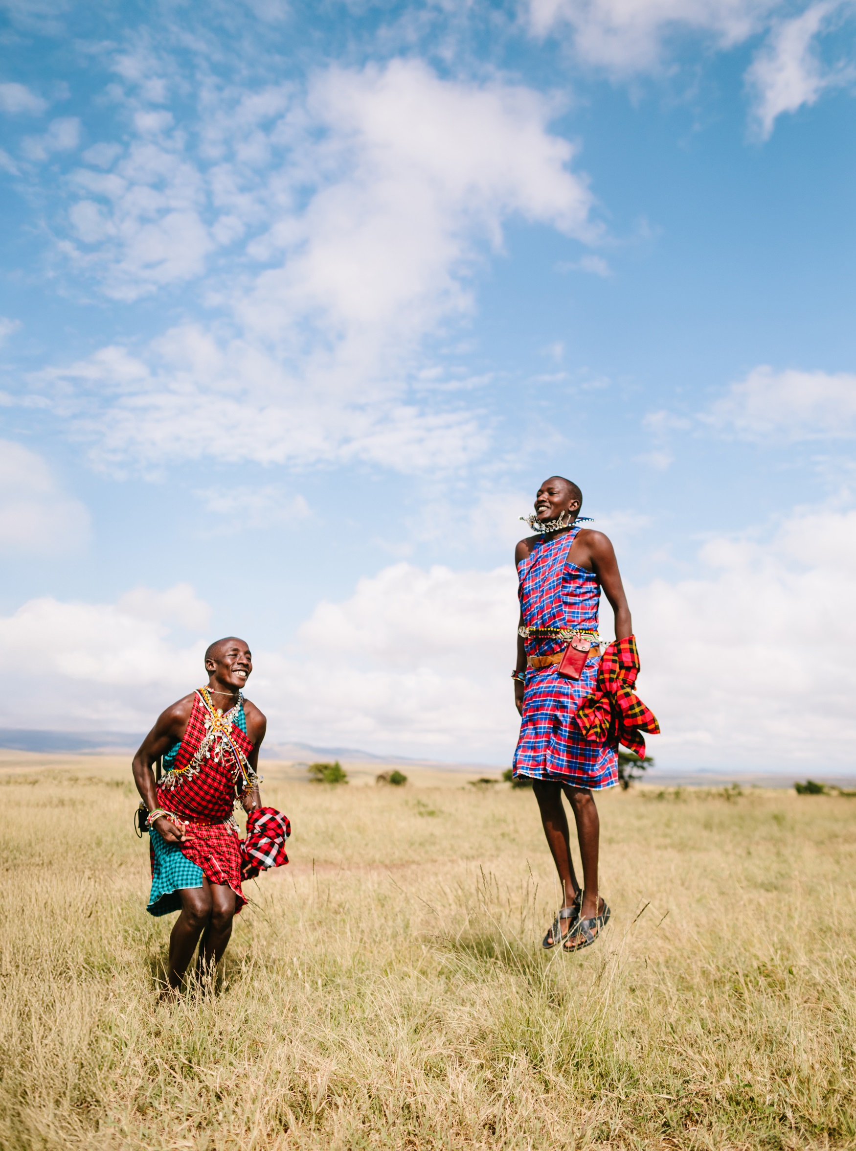 Two Maasai men in colorful traditional attire smile joyfully while jumping high in a grassy field under a blue sky, sharing their culture with families visiting the Kenyan savannah.
