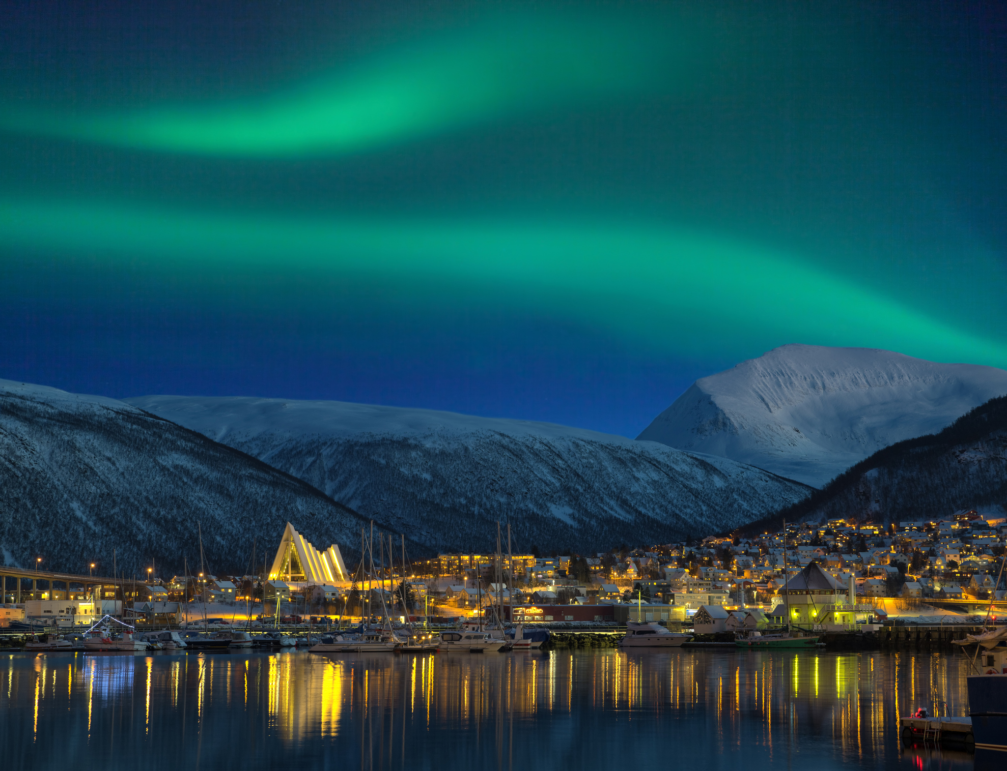 A vibrant aurora borealis over a snow-covered mountainous landscape with illuminated buildings and boats along the waterfront at night.