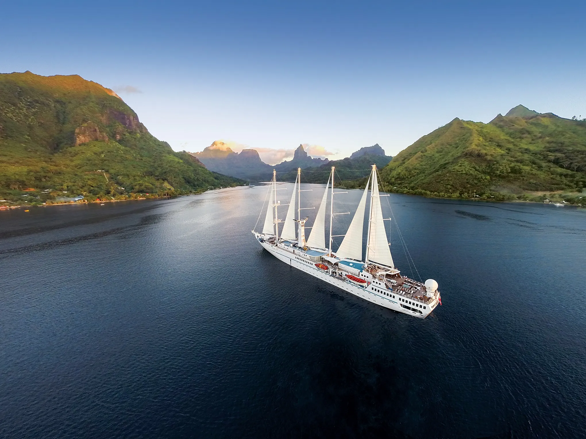 A large white sailing ship with multiple masts cruising through calm water, surrounded by lush green mountains under a clear blue sky.