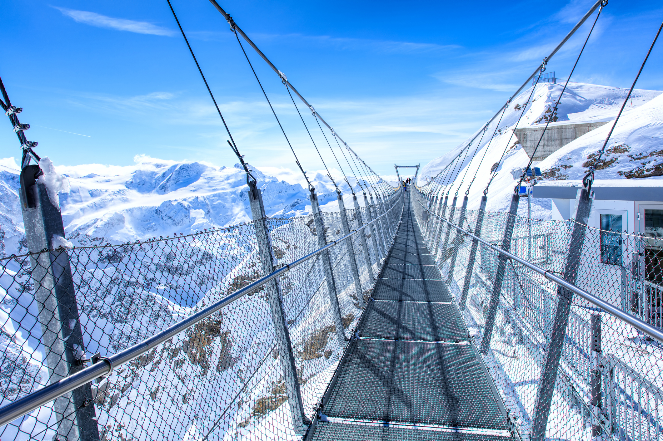 The Titlis suspension bridge with a metal mesh floor and side fencing spans across a snowy mountain landscape under a clear blue sky.