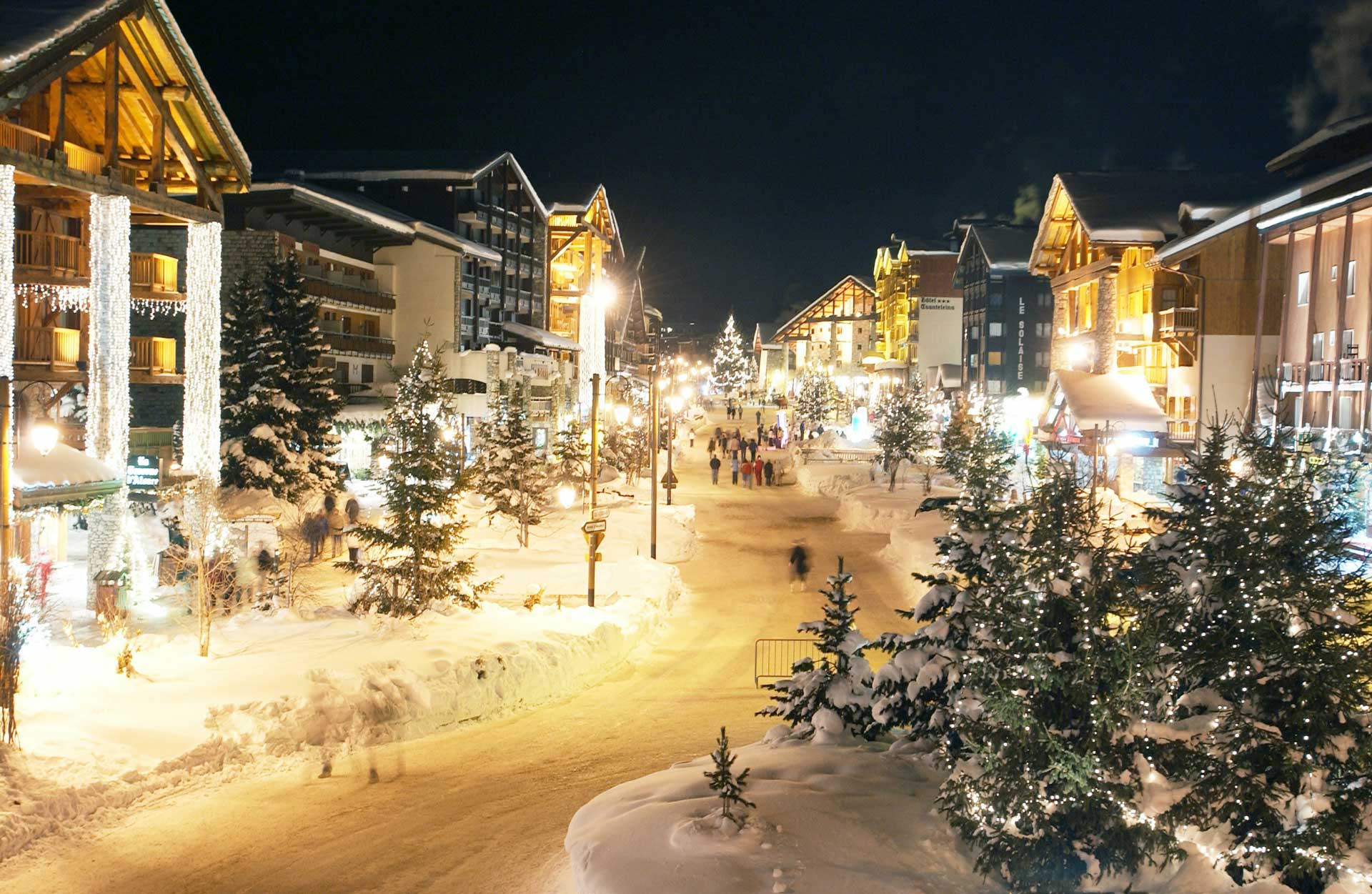 The centre of a winter village with people walking a flattened pathway lit up at night