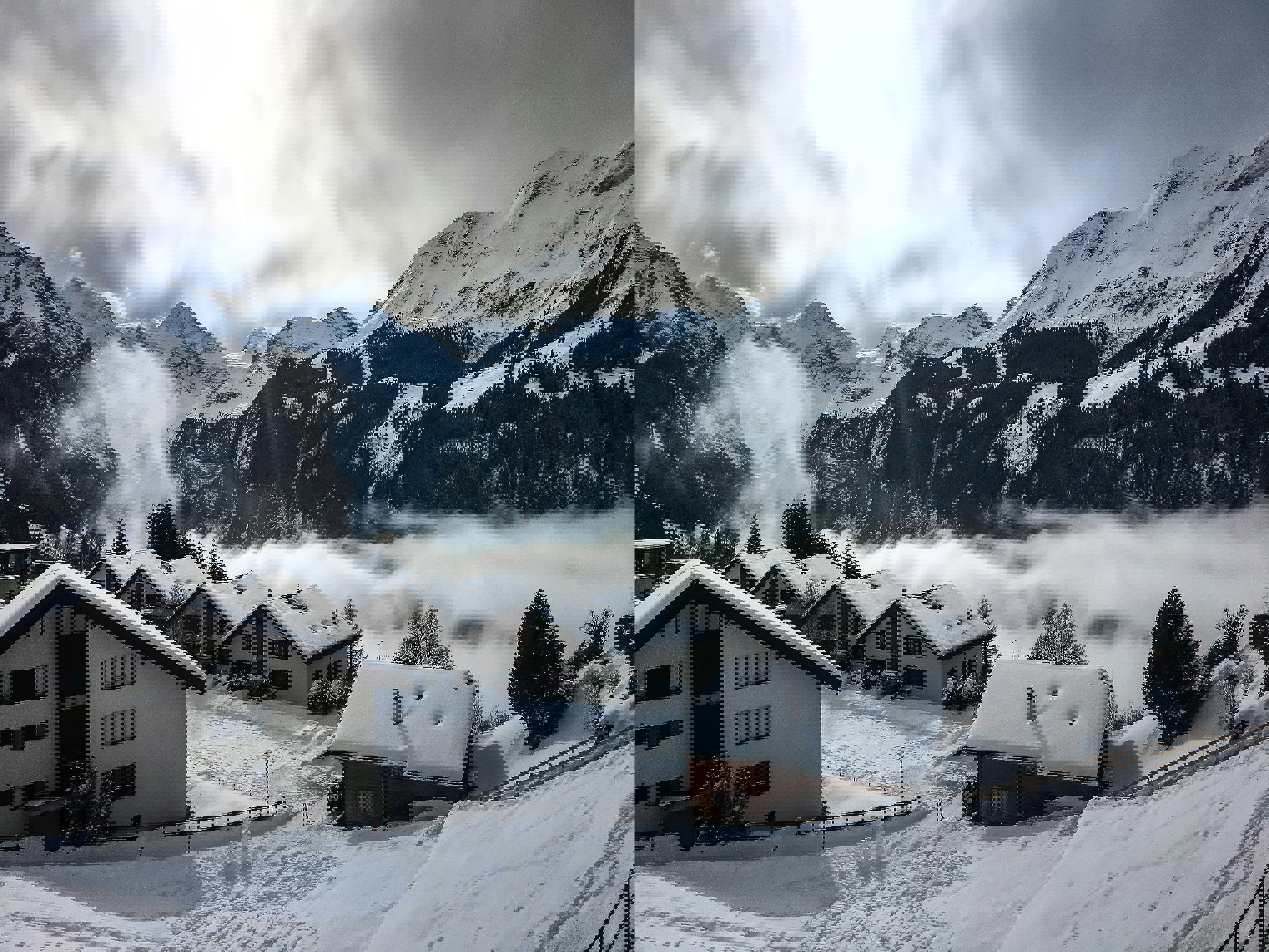 A snowy scene of Wengen in Switzerland with snow-covered buildings in front of white mountains with the sun breaking through the clouds