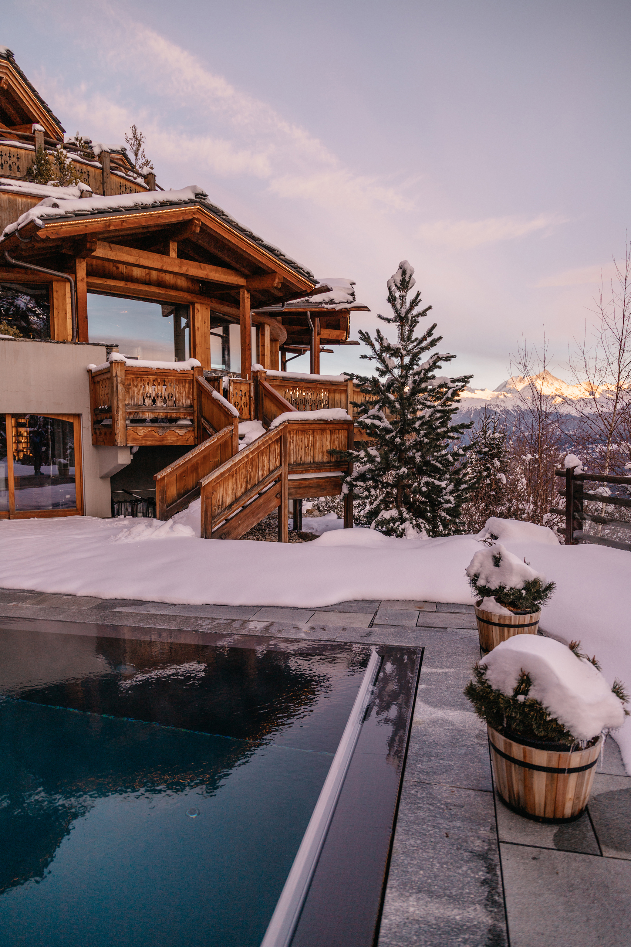A wooden lodge and stairs leading to an outdoor swimming pool with plant pots on a terrace covered in snow