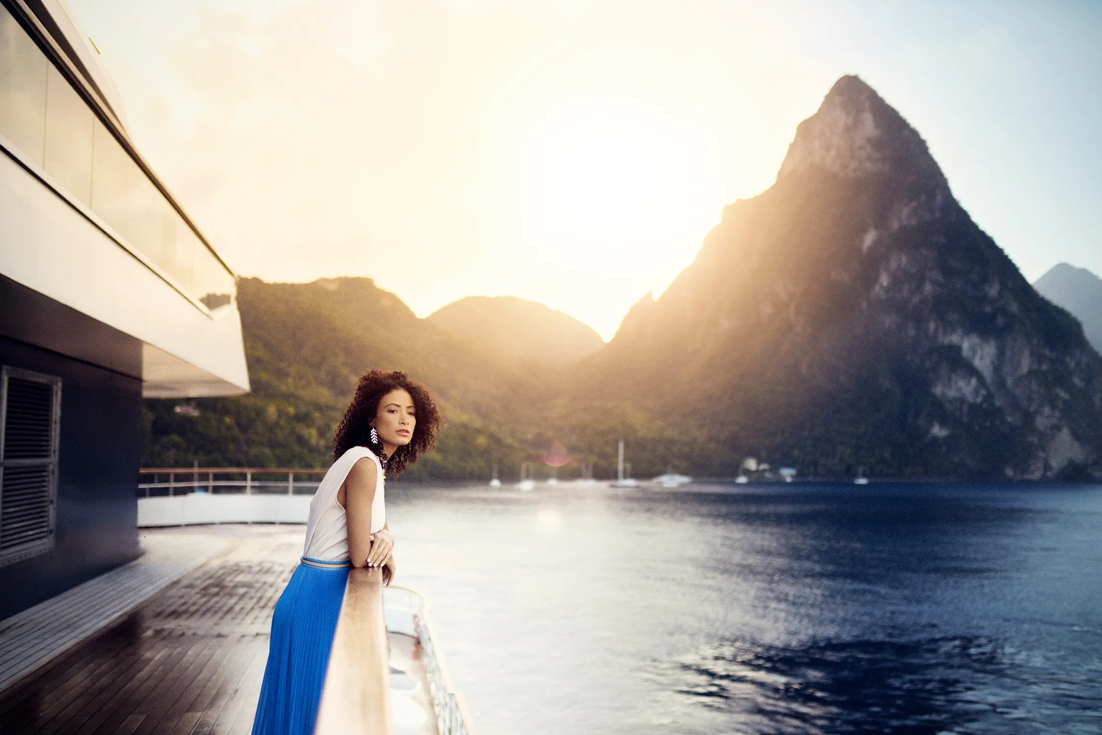 A woman stands on a boat deck at sunset, gazing at mountains and water in the distance.