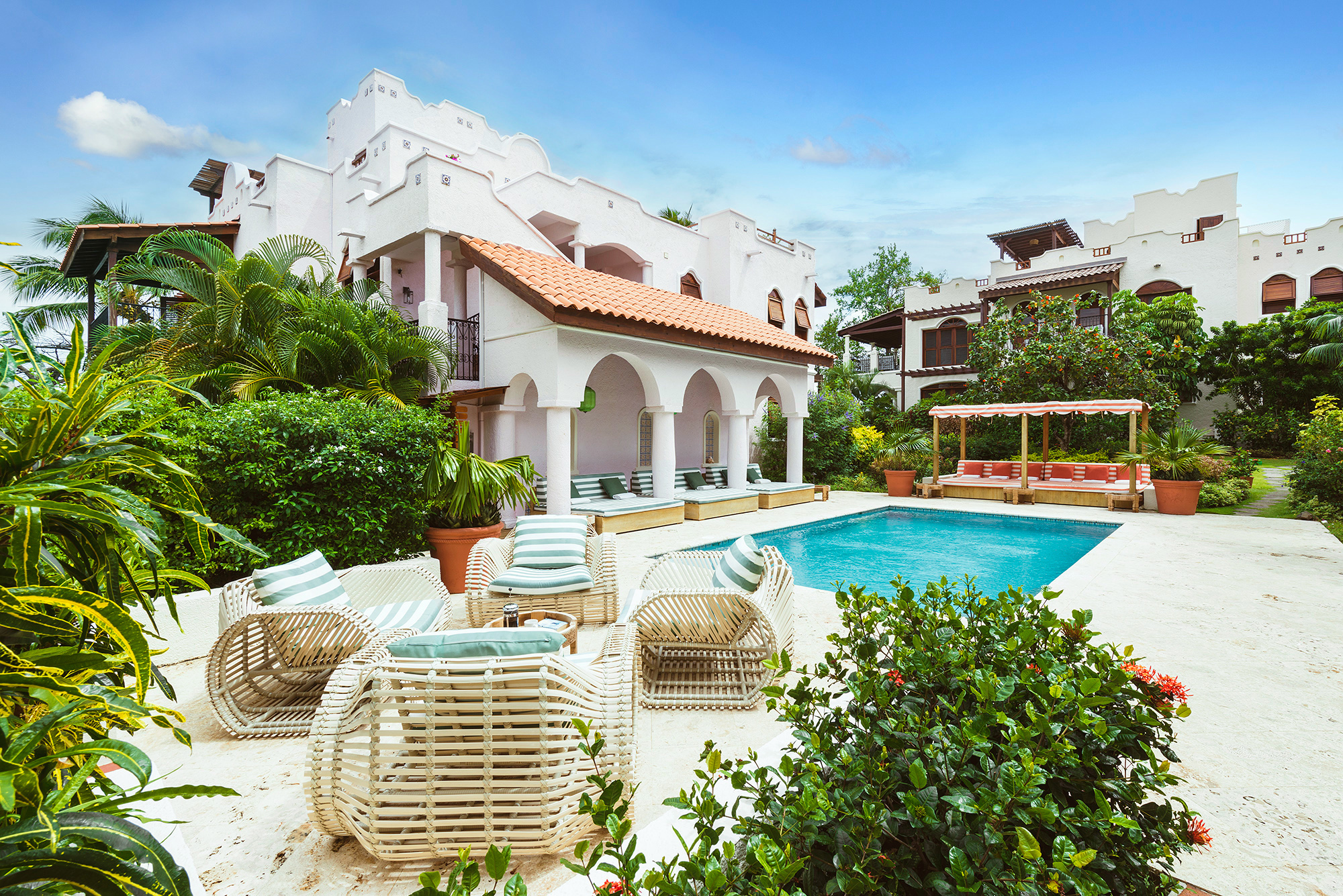 A large pool courtyard outside a whitewashed building at Cap Maison featuring a pool, white rattan furniture and lush greenery