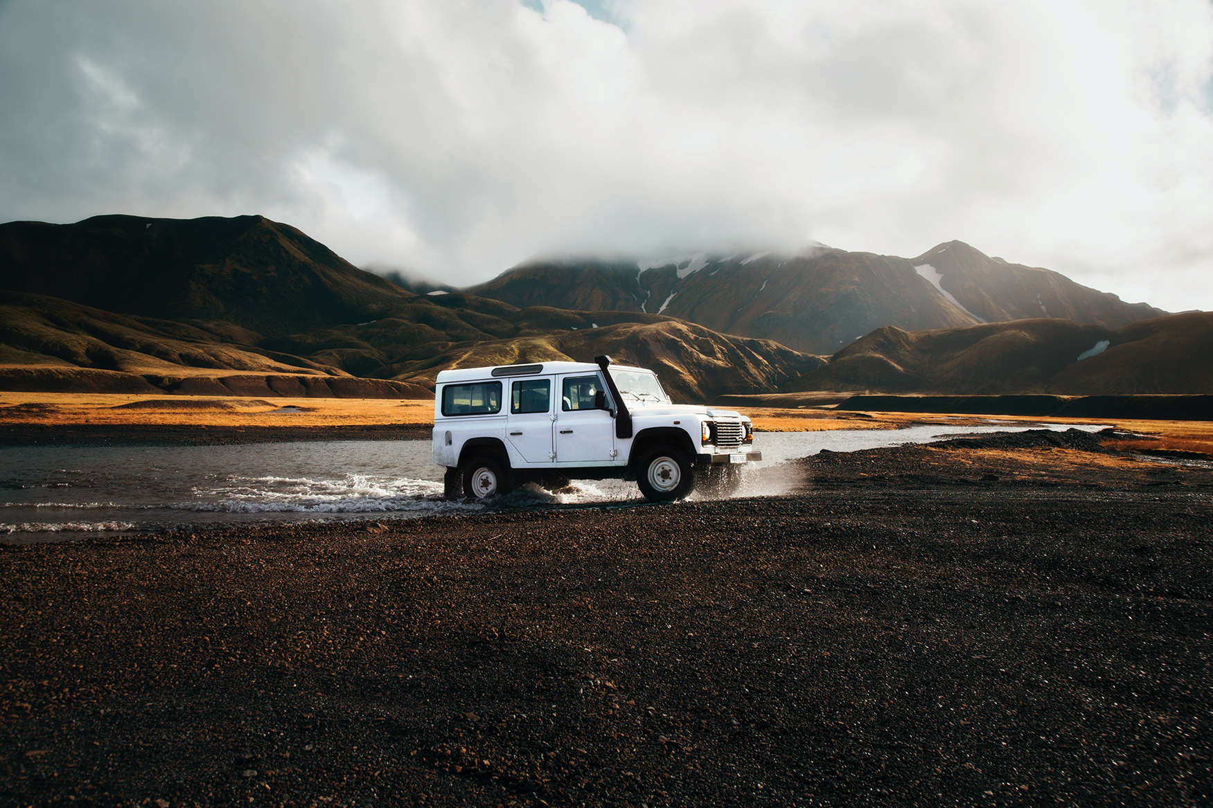 A white SUV exiting water onto black sand with the mountains of Landmannalaugar, Iceland in the background