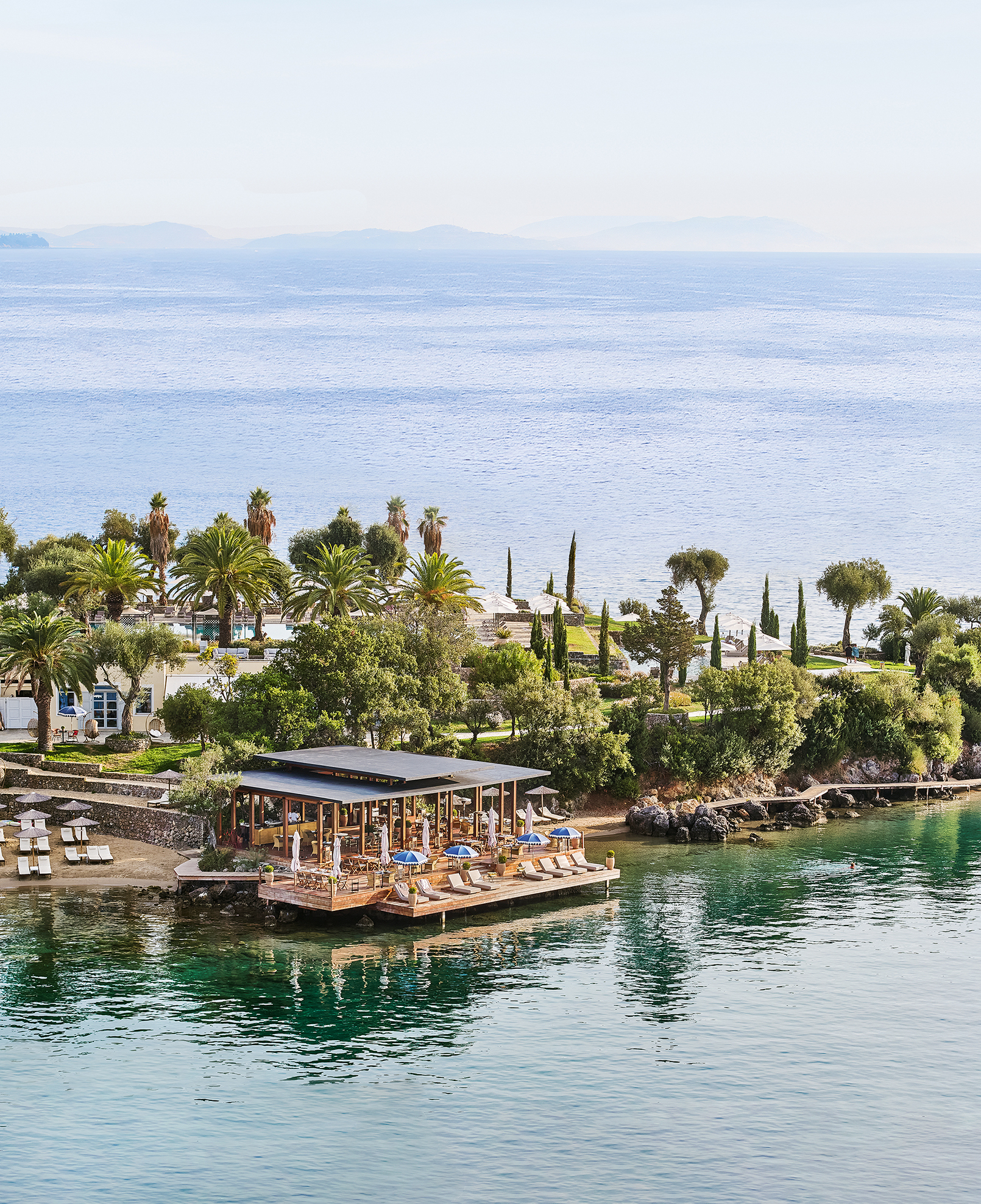 Aerial view of Yali Seafood Restaurant at Grecotel Corfu Imperial showcasing lush gardens, palm-lined waterfront, and overwater dining area.