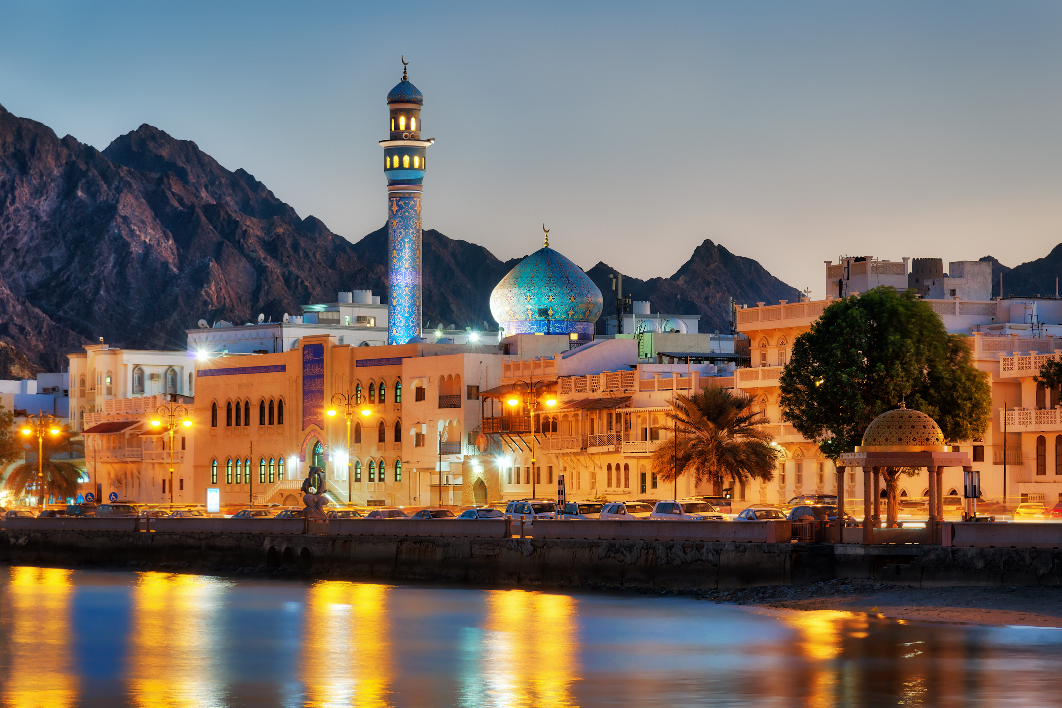 A scenic view of Muscat, Oman at dusk featuring a mosque with a blue dome and minaret, surrounded by traditional buildings and mountains in the background