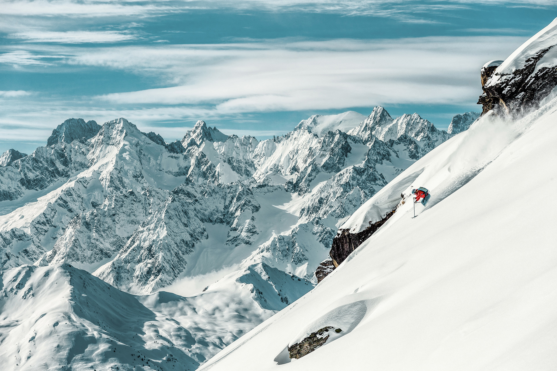 A skier going down a sharp snowy incline with rocky mountains in the background