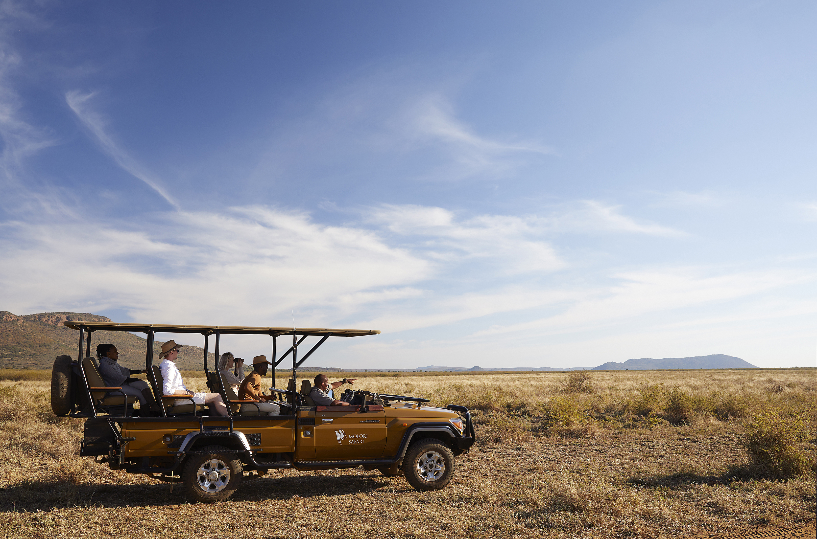 A brown safari jeep parked in the middle of the grassy savannah with rangers and guests inside beneath a clear blue sky