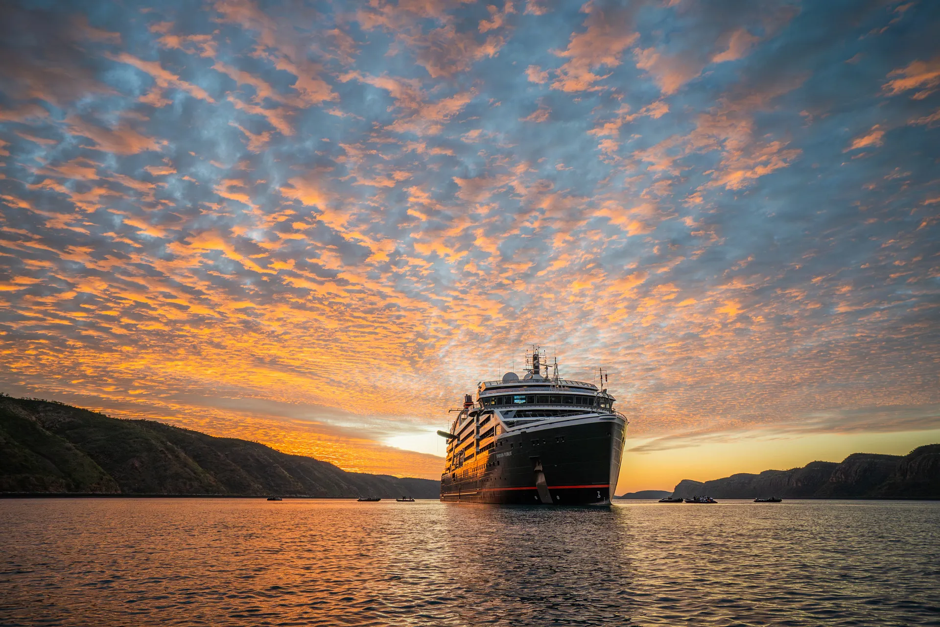 Cruise ship sailing on calm waters beneath a vibrant sunset sky with scattered clouds and distant hills.