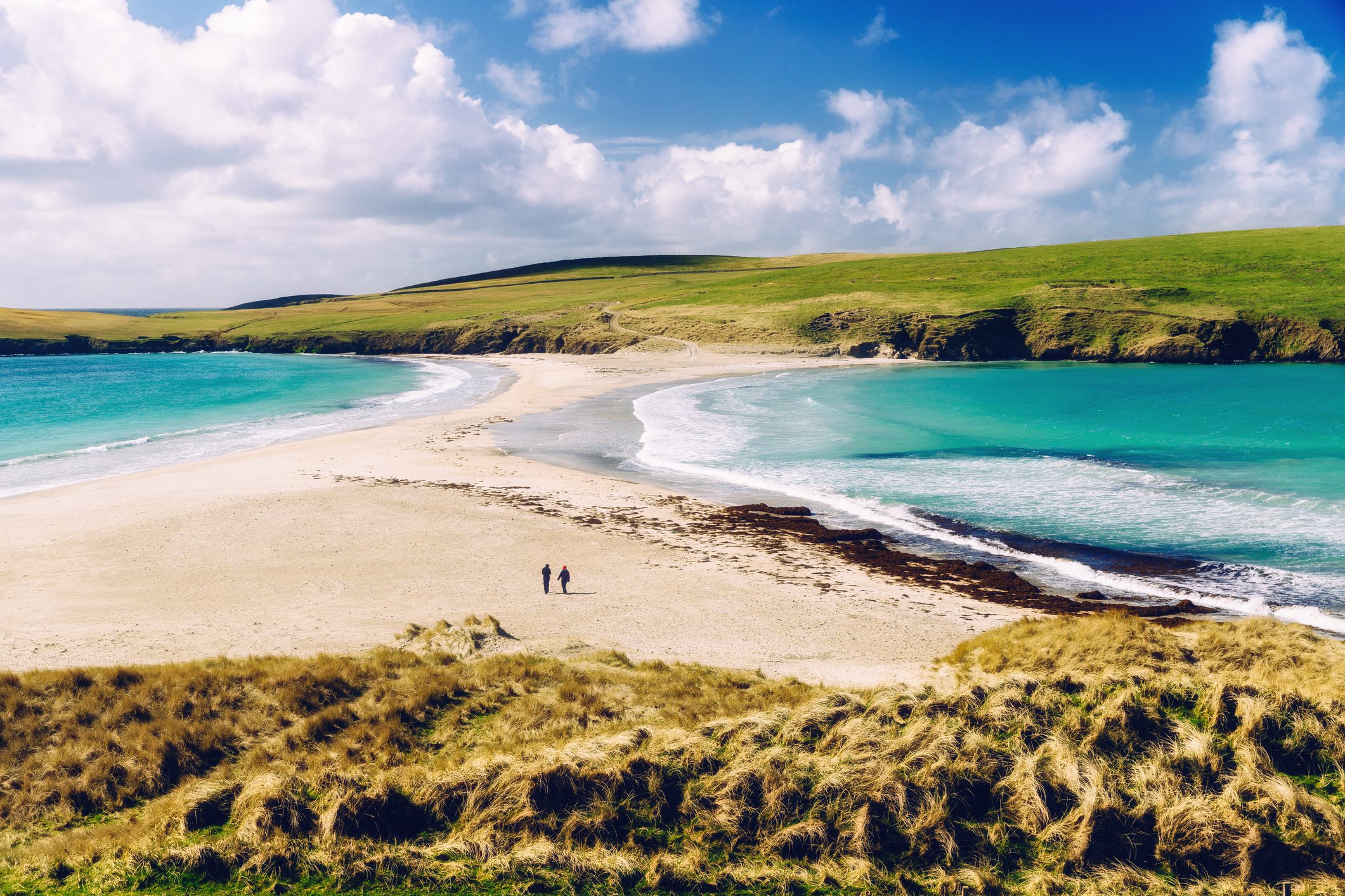 A panoramic view of a serene beach with turquoise waters, green hills, and two people walking on the sand under a blue sky with scattered clouds.