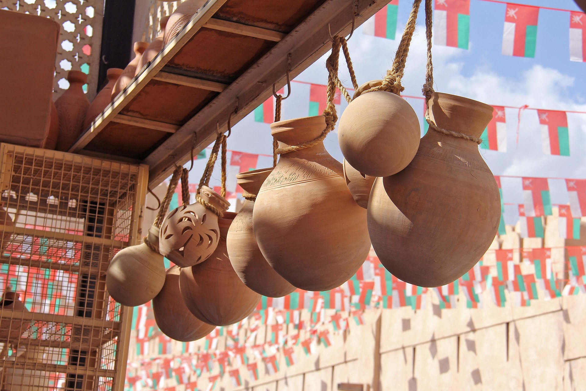 Clay pots hanging from ropes under a wooden structure with colorful flags in the background in a souk in Nizwa, Oman