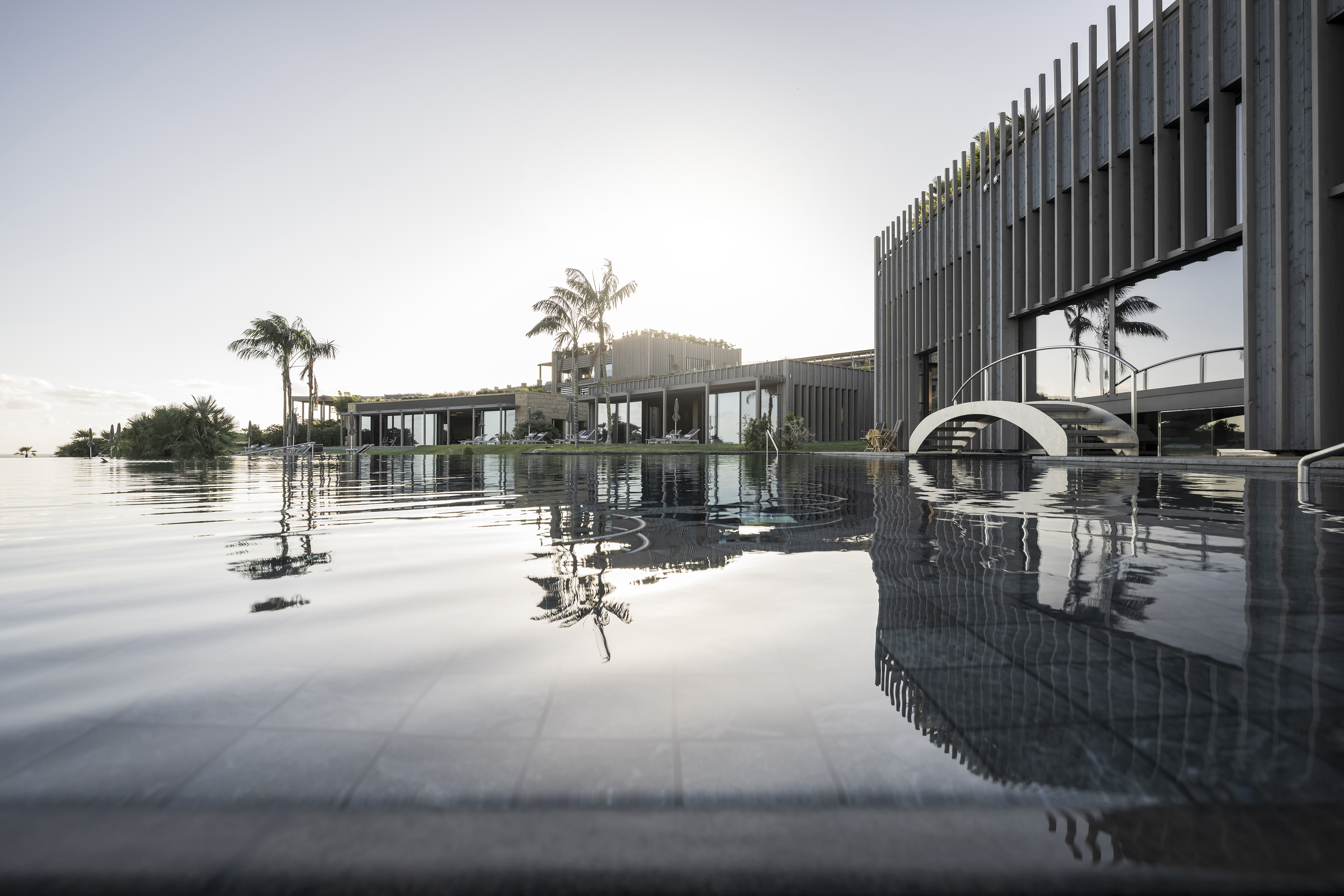 A vast infinity pool set in front of a wood-faced building and bridge flanked by palm trees at sunset