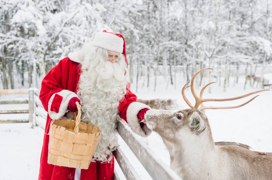 A person dressed as Santa Claus feeding a reindeer in a snowy setting.