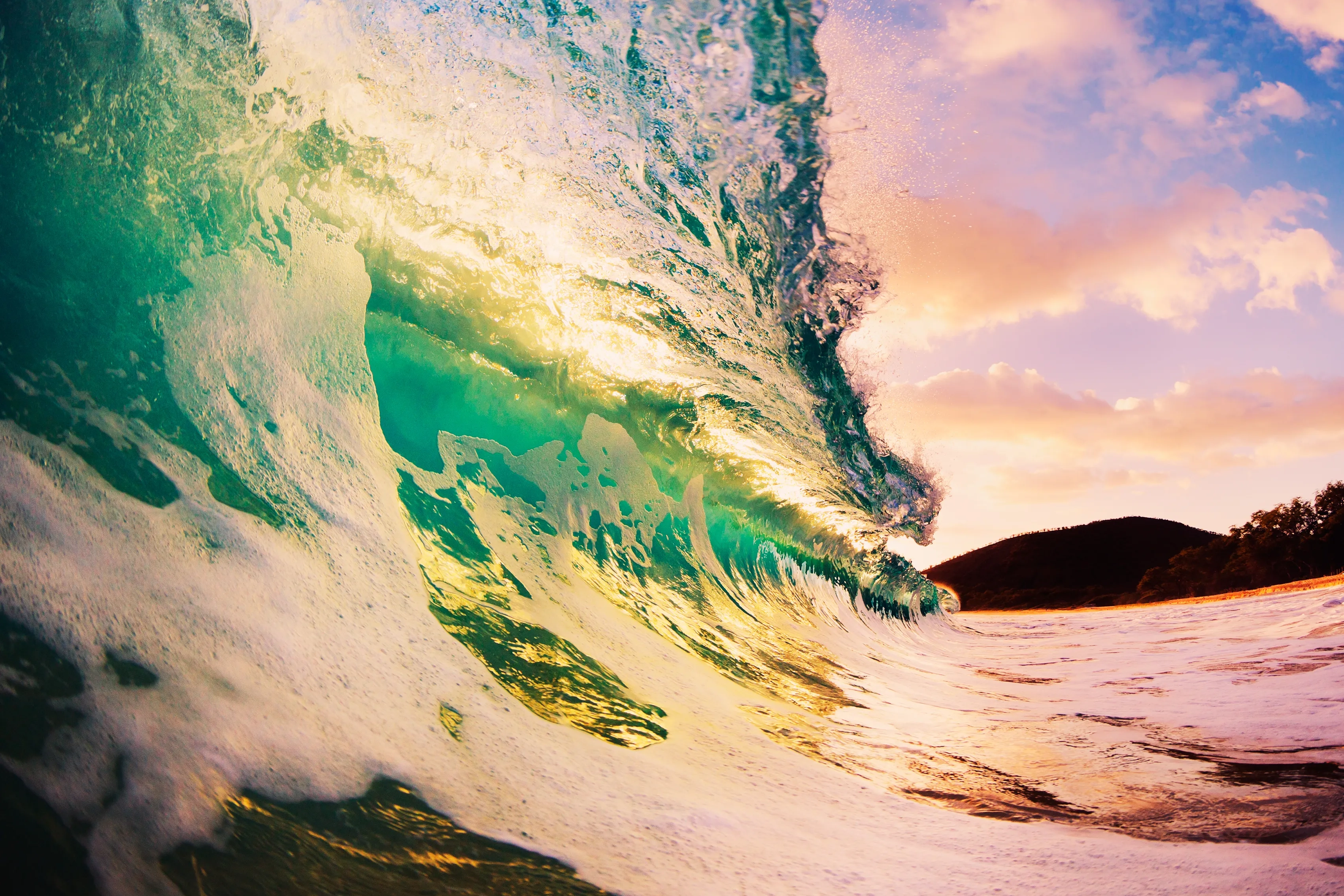 A clear wave about to break overhead lit up with green, pink and blue at sunset, with a glimpse of land behind