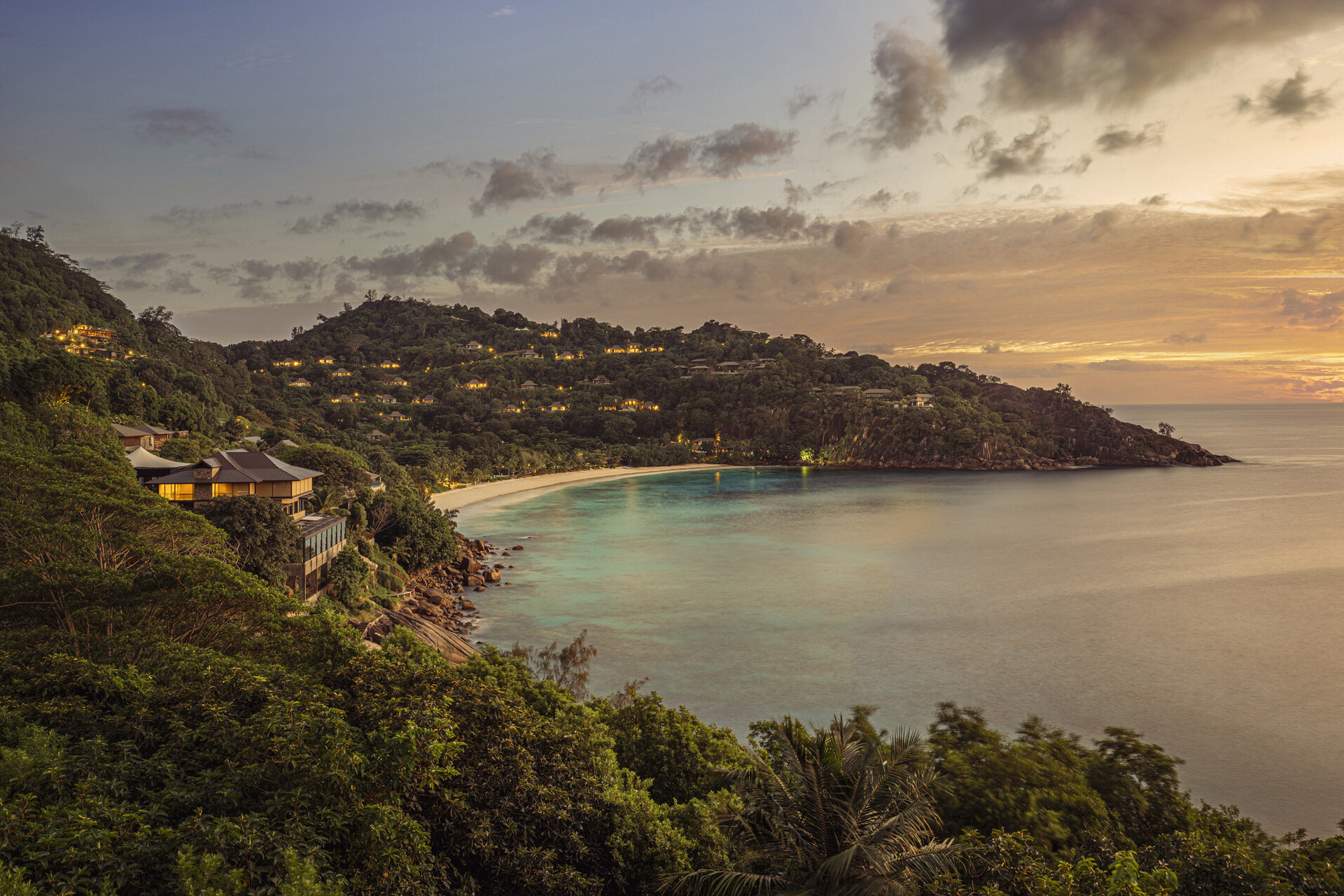 Lush green crescent bay of Four Seasons Seychelles with sandy beach and lights on in the villas