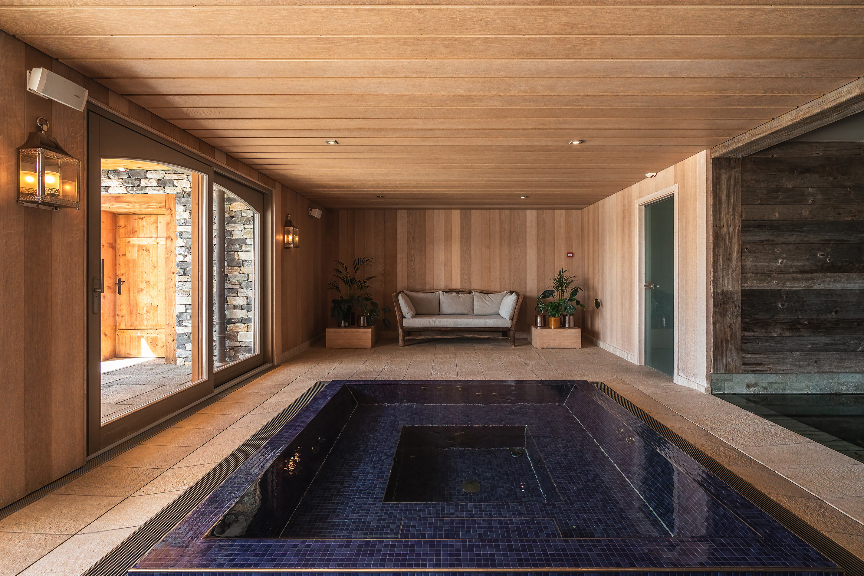 A blue square plunge pool in the middle of a pale wooden spa room with a sofa and glass walls looking into other rooms