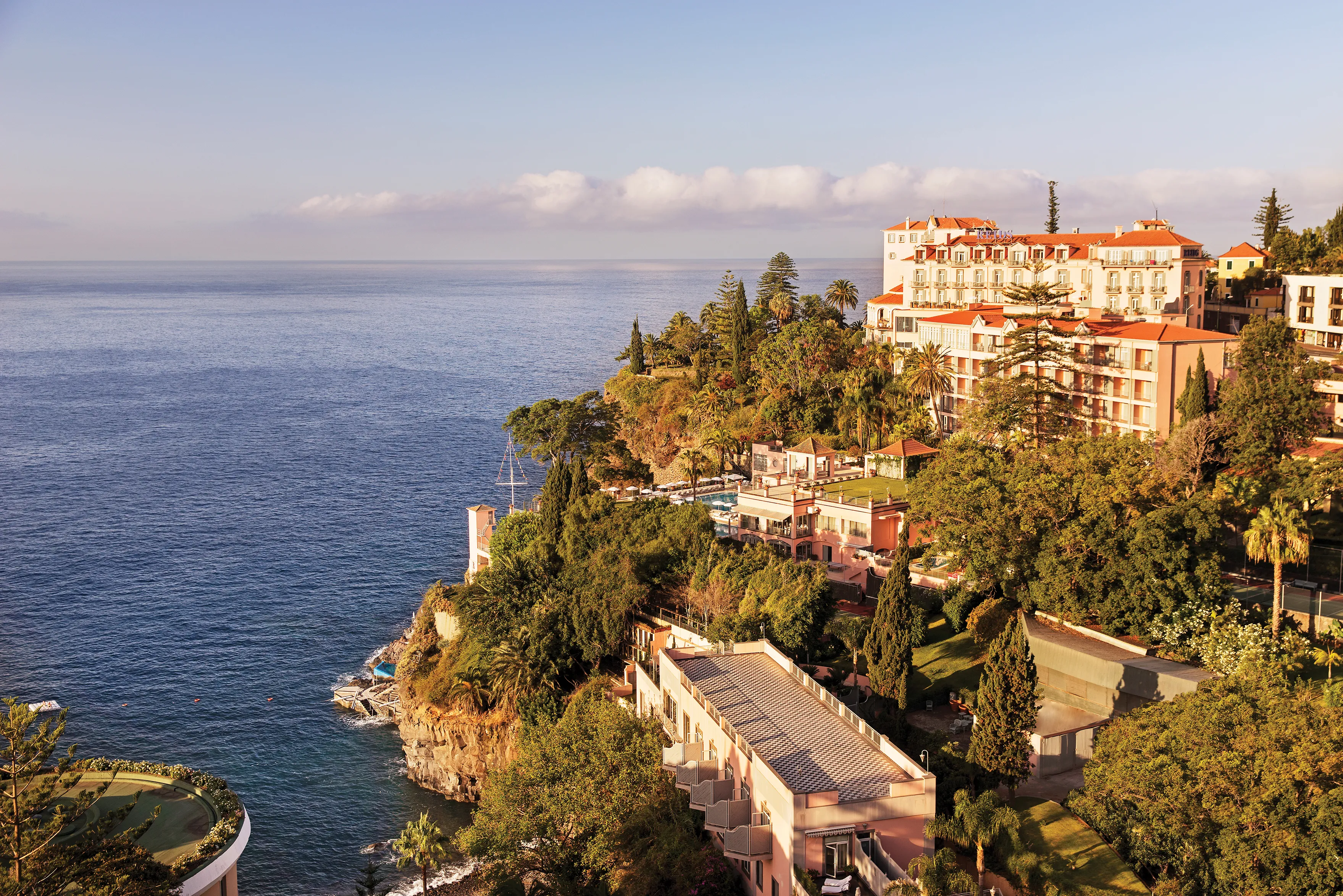 Cliffside view of Reid’s Palace, A Belmond Hotel, set among lush gardens overlooking the Atlantic Ocean with dramatic coastal scenery and sunlit terraces.
