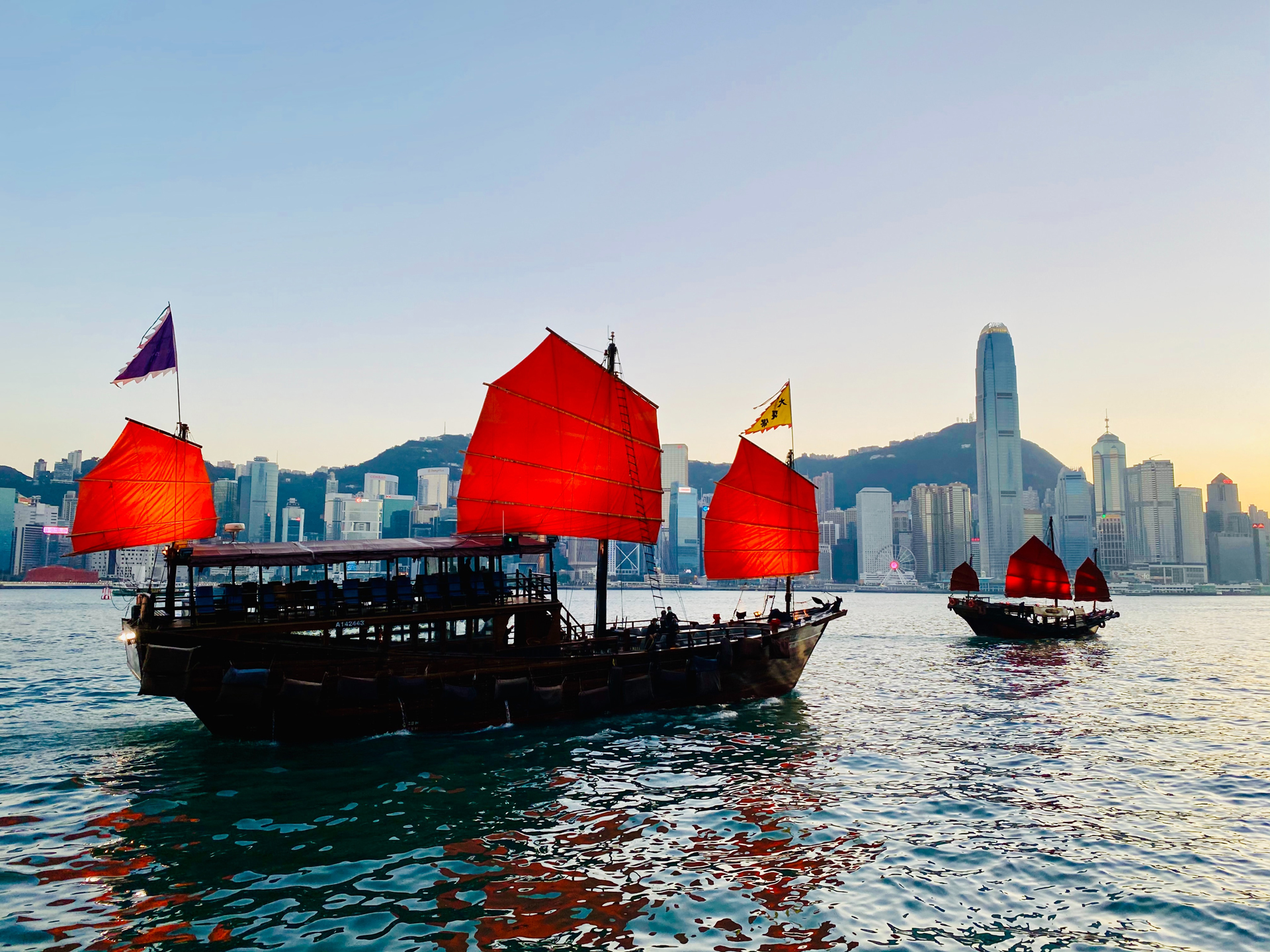 Two boats with red sails on water with a city in the background