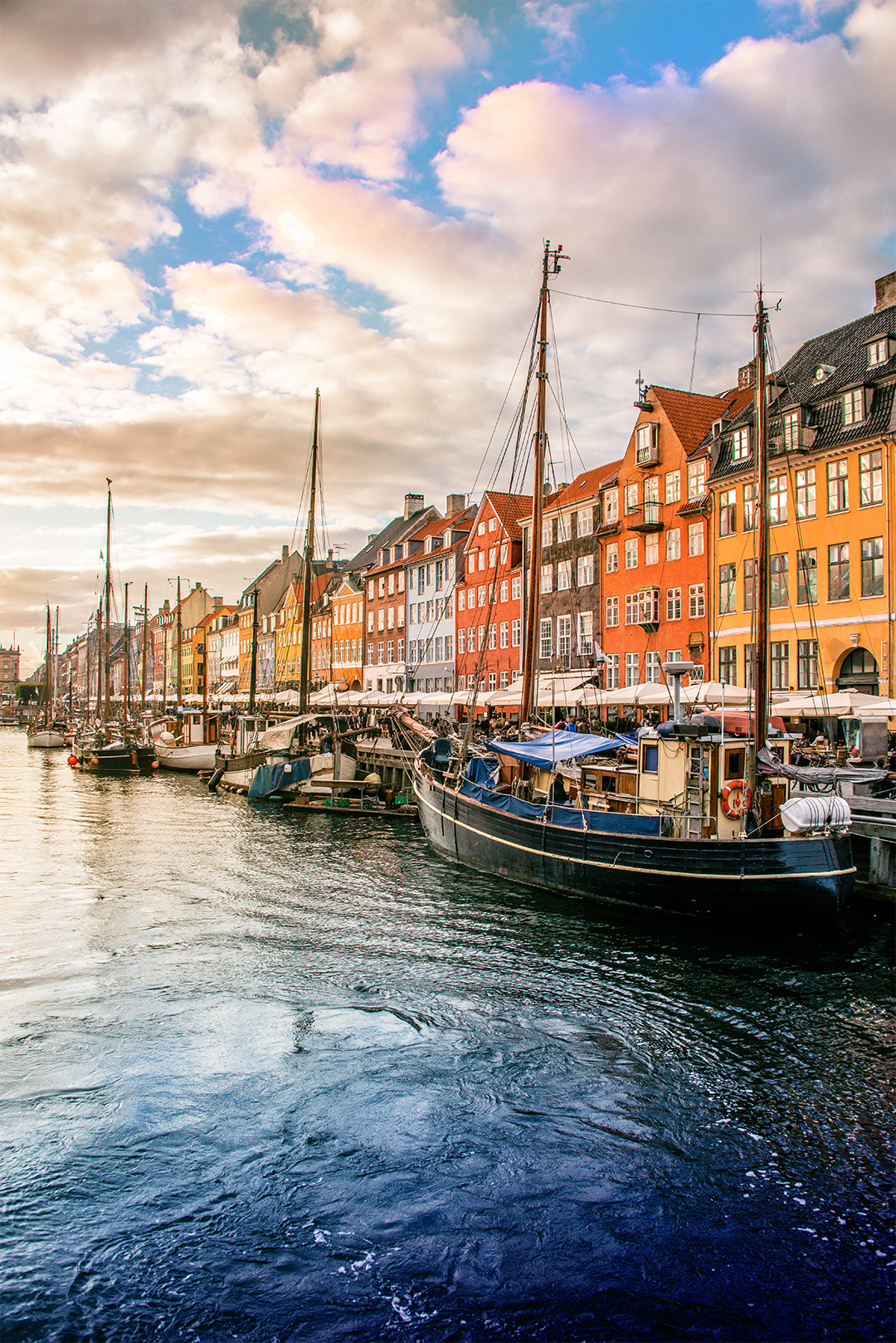 A scenic view of a canal with boats moored alongside colourful buildings under a partly cloudy sky at sunset.