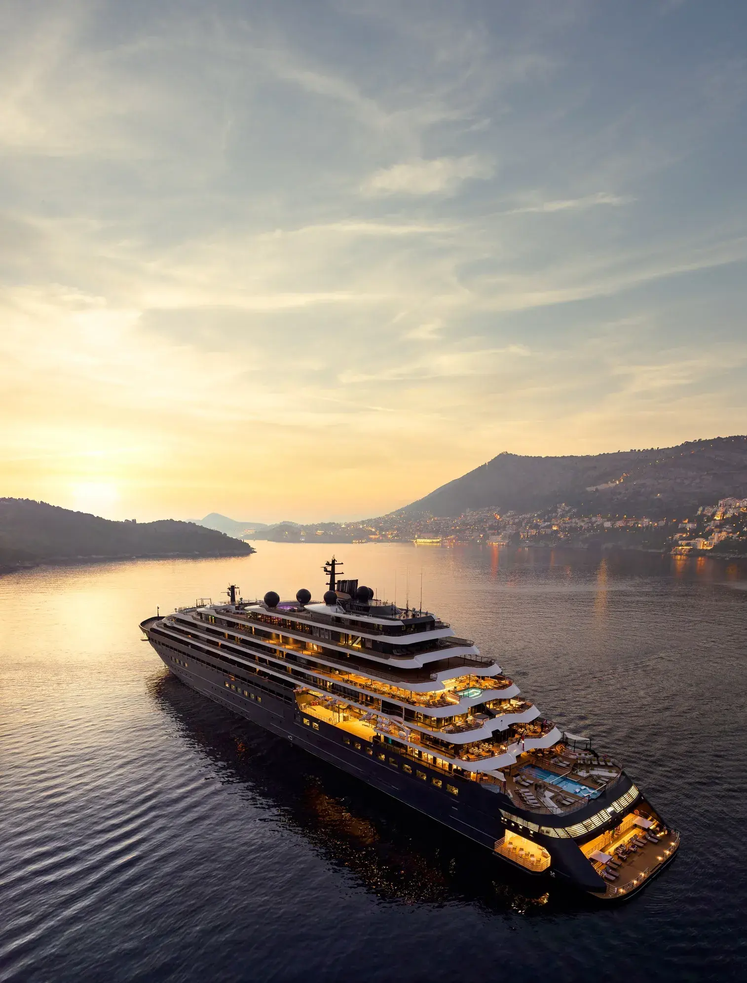 A large luxury yacht sailing through calm waters at sunset near hills and a distant town