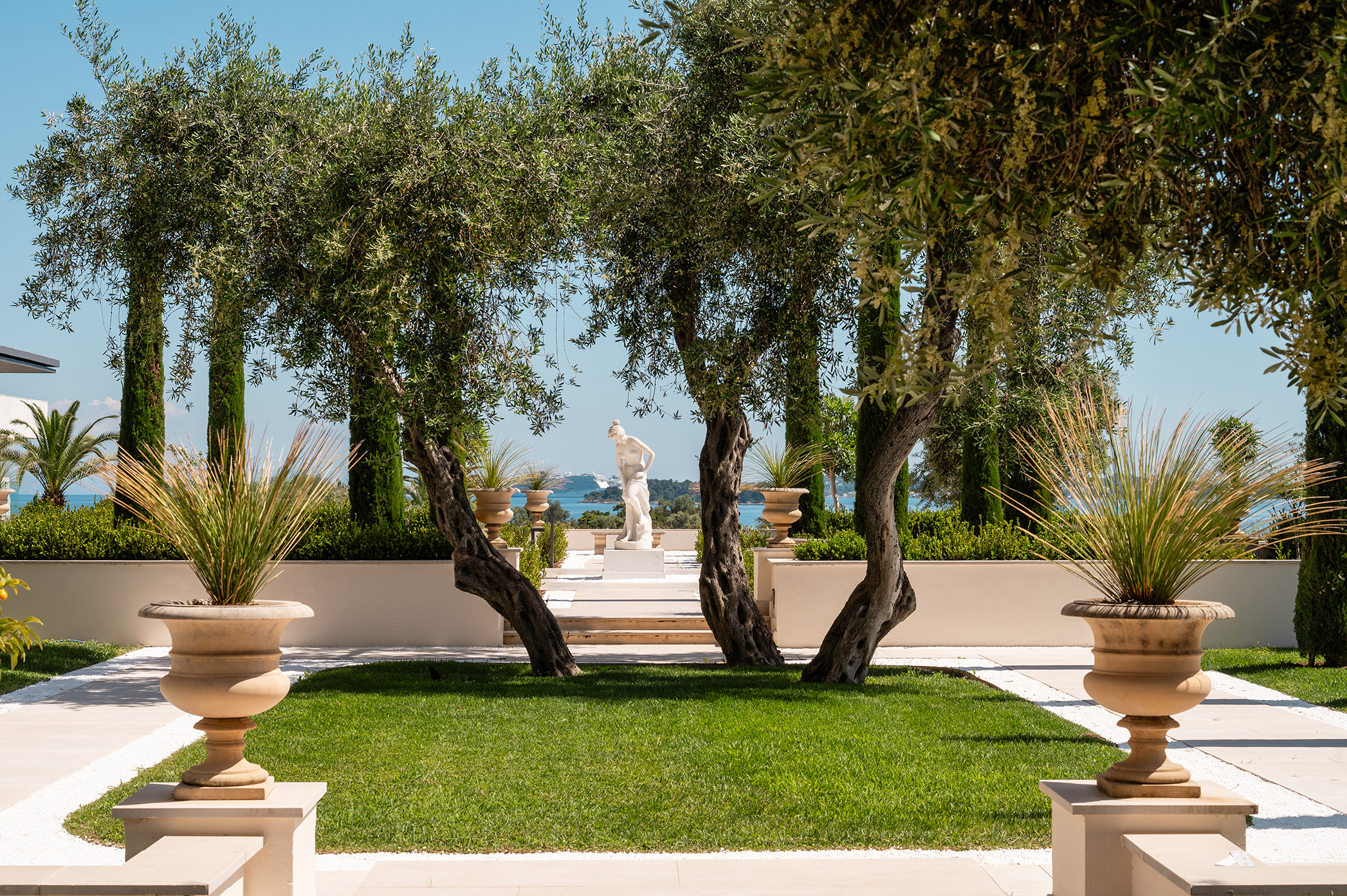 Elegant garden view at Grecotel Corfu Imperial featuring lush greenery, classical statues, and a serene sea backdrop.