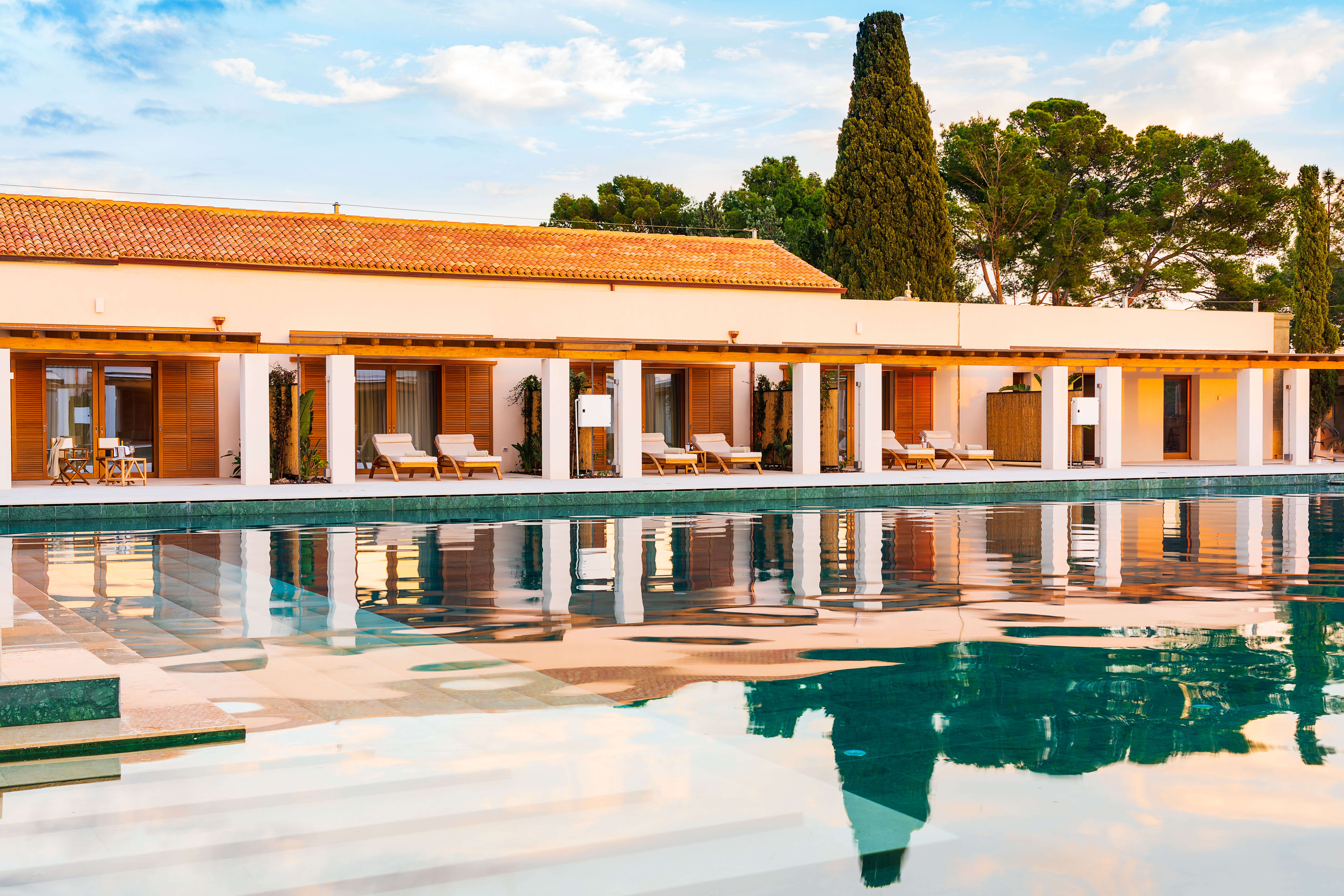 A calm pool in front of a terracotta building at Il San Corrado with sun loungers outside and trees poking out from above the roof