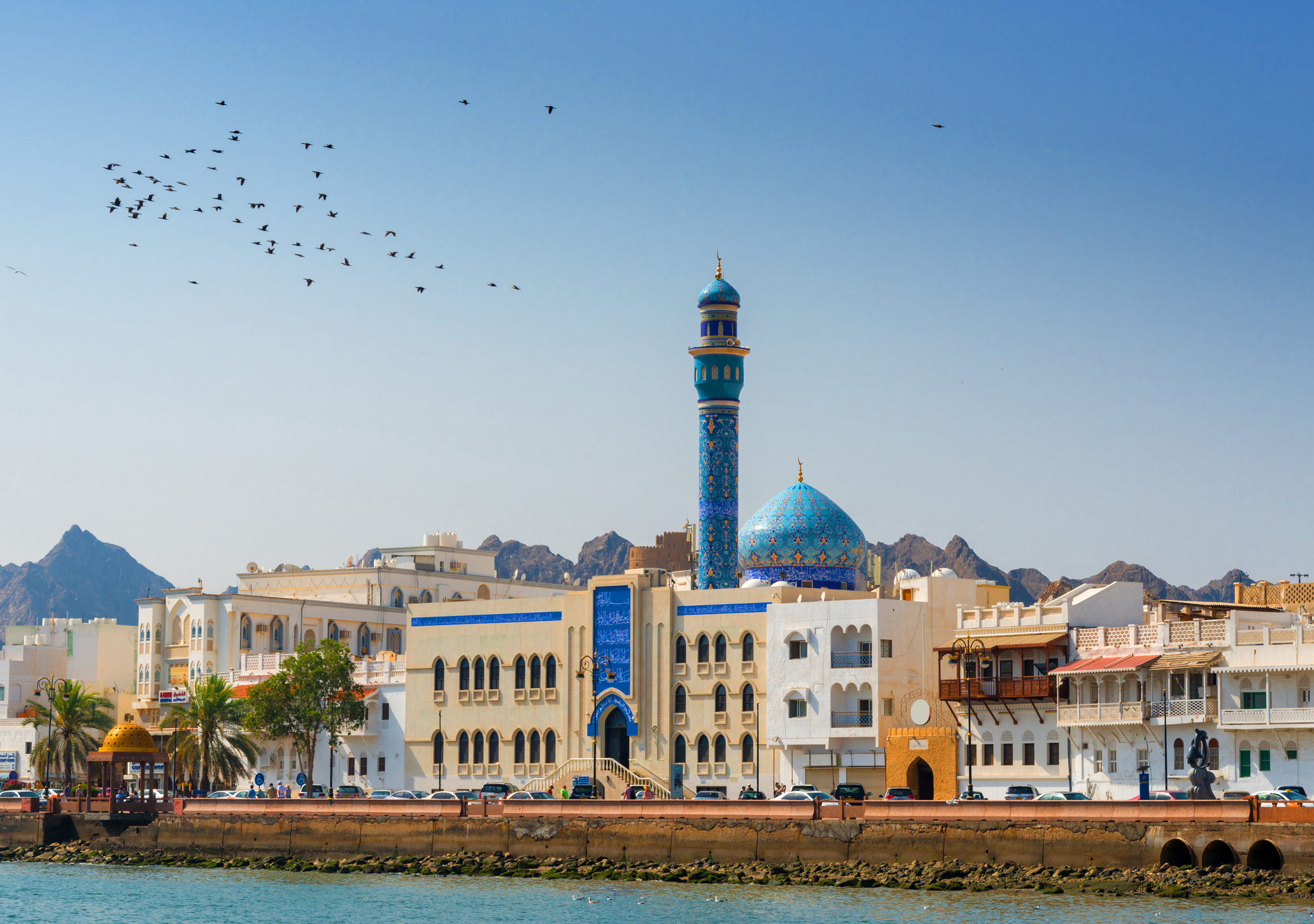 A coastal cityscape with traditional white buildings, a prominent blue and gold patterned mosque minaret, under a clear sky with birds flying and mountains in the background.