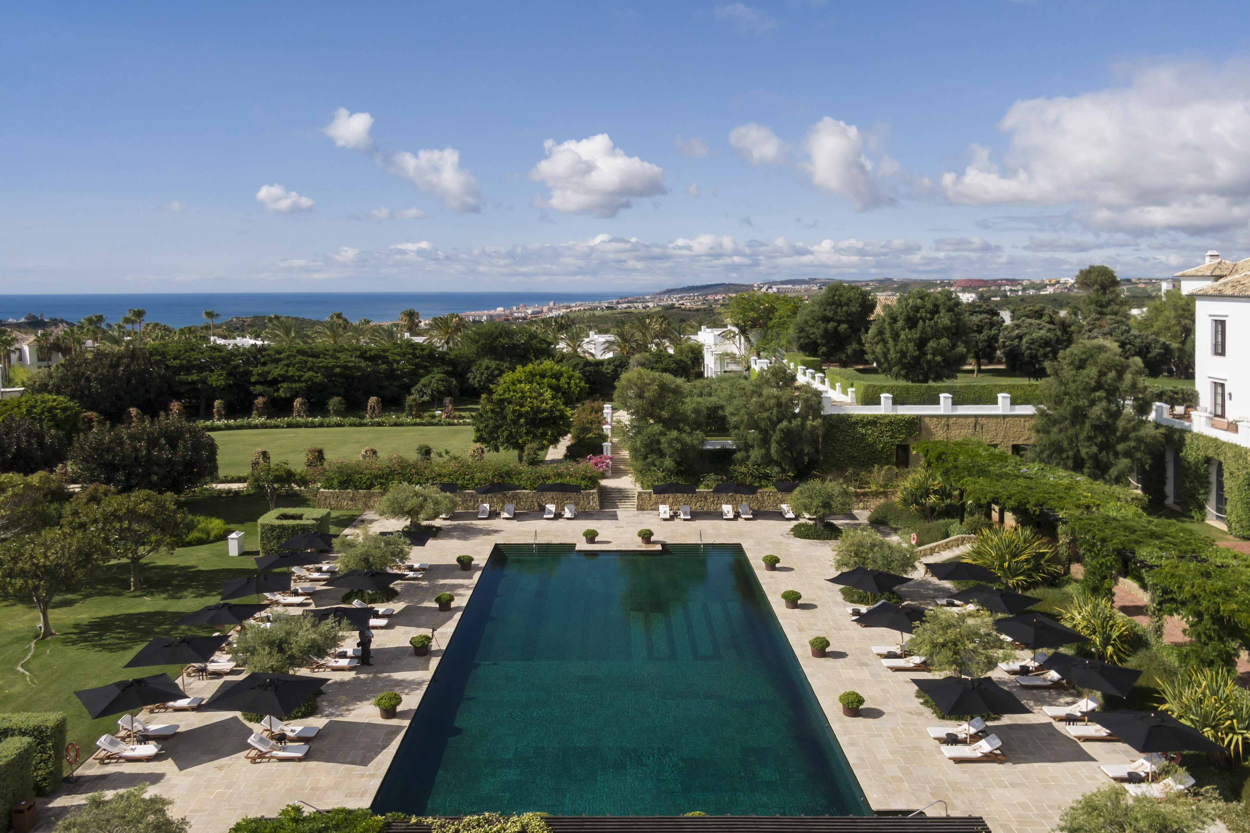 The large swimming pool of Finca Cortesin under a clear sky, bordered by trees and comfortable seating areas for guests