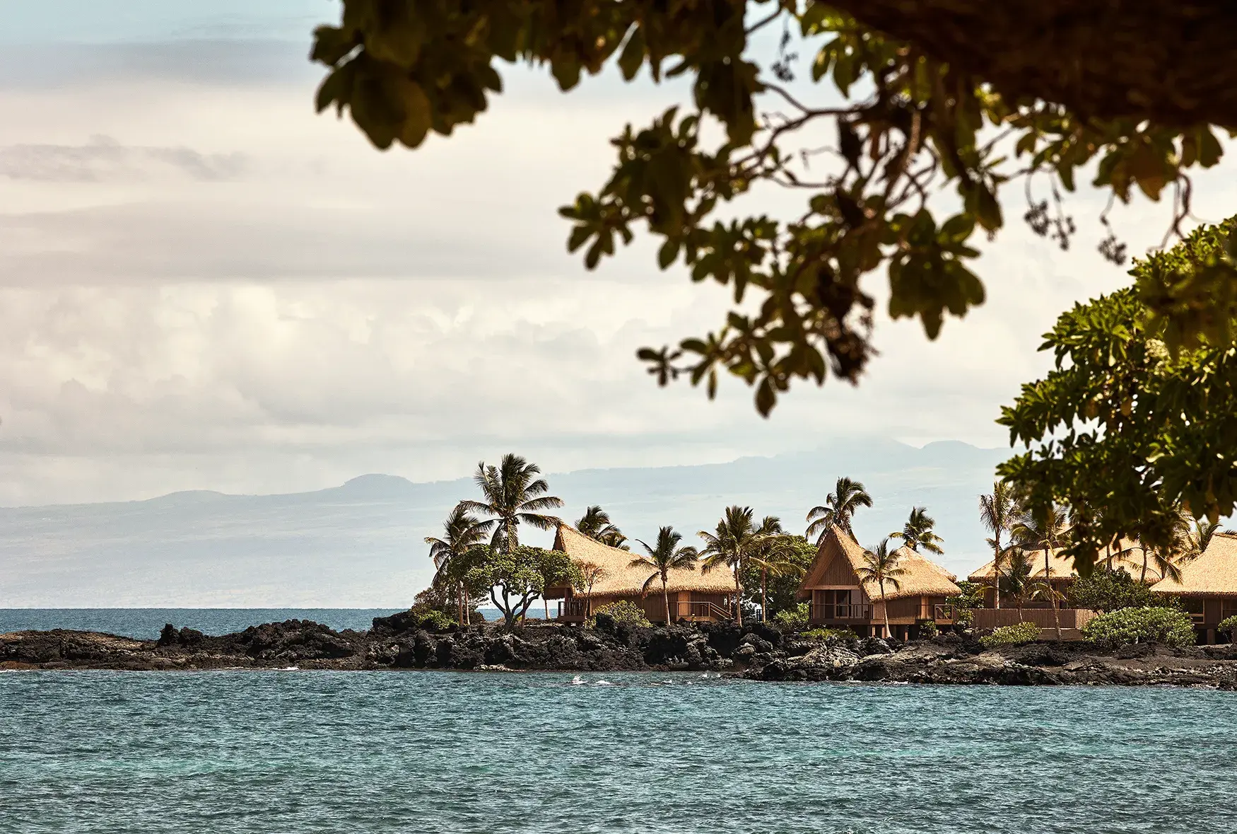 Thatched hale accommodation at Rosewood Kona Village perched along the coastline among palms with misty mountains in the background