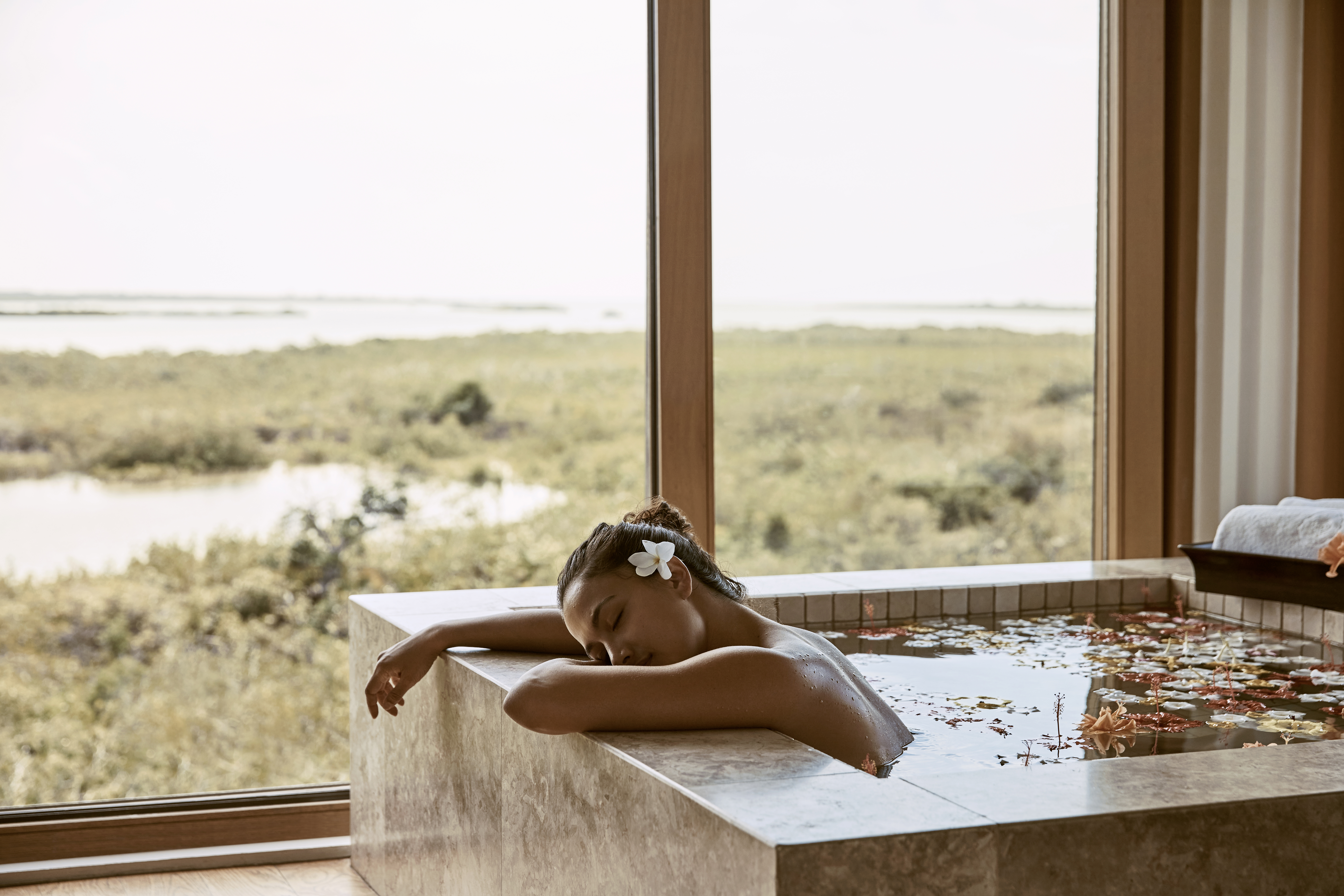 A relaxed woman lounging on the side of a deep bath in front of a large window at COMO Parrot Cay