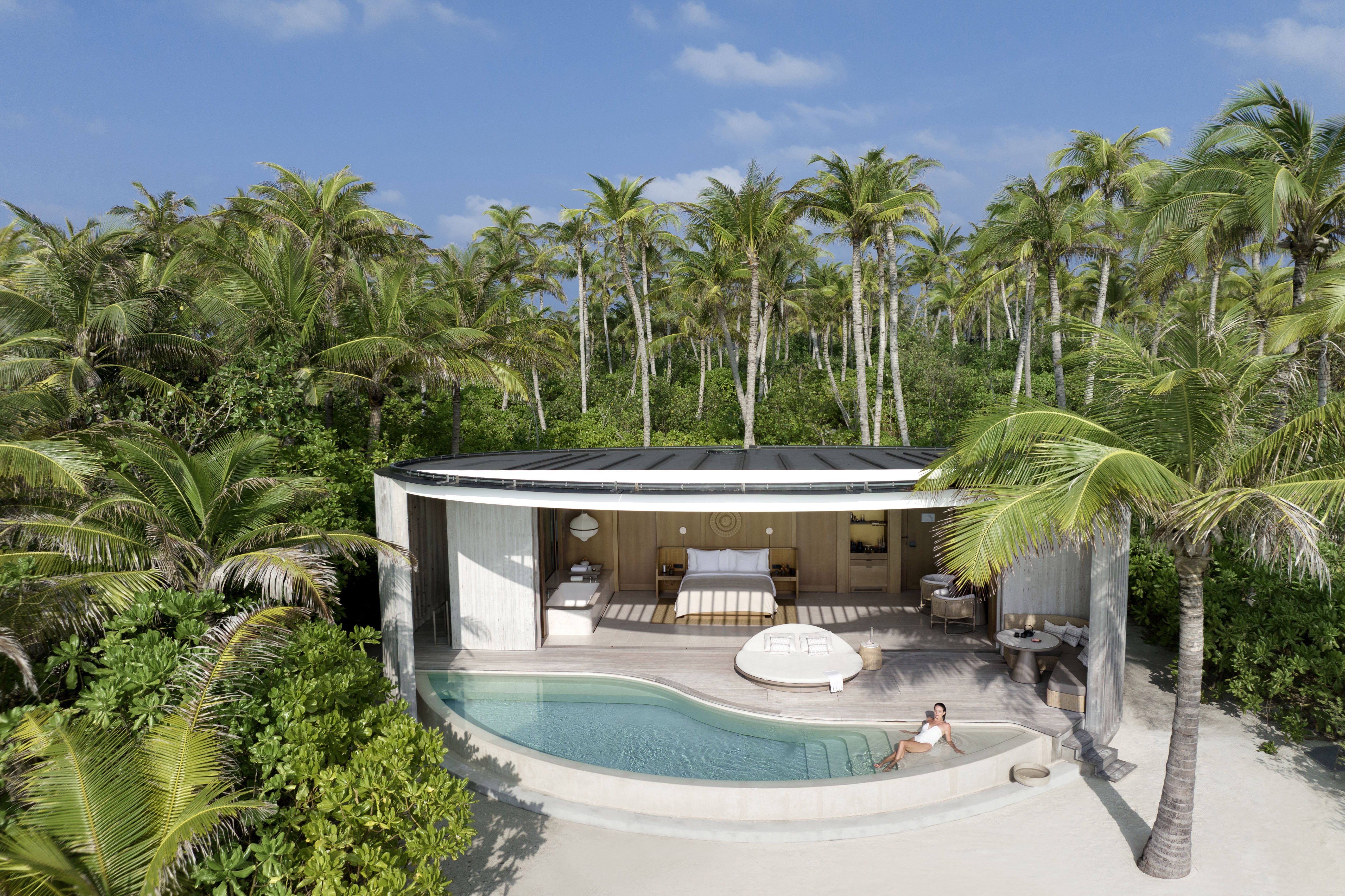 A woman lounging in the private pool of a villa at The Ritz-Carlton Maldives, Fari Islands, with a bedroom featuring a large bed and sun lounger behind