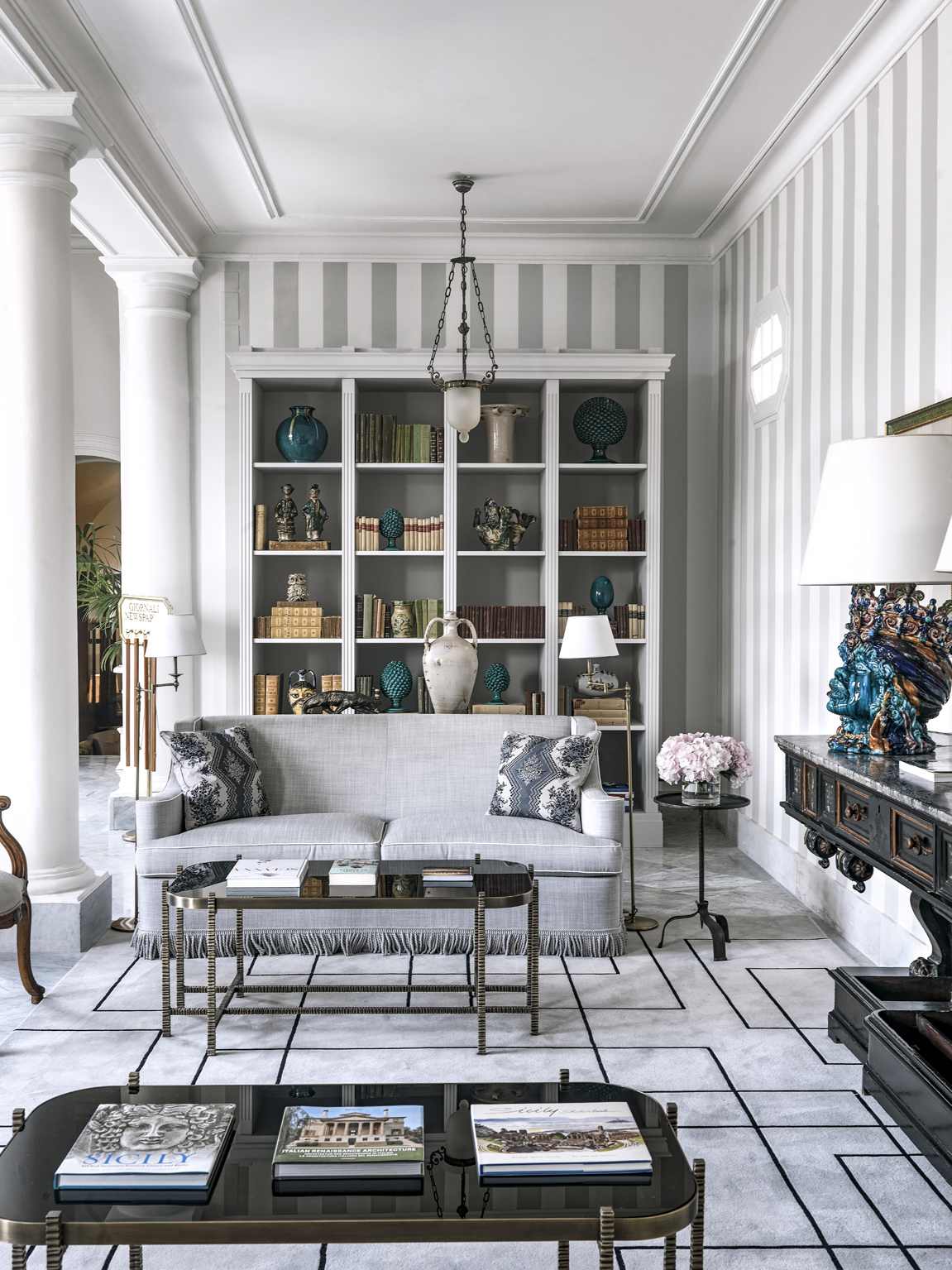 An elegant library with a white and gray colour scheme, featuring striped sofas, a chandelier, bookshelves with decorative items, and a classic fireplace.