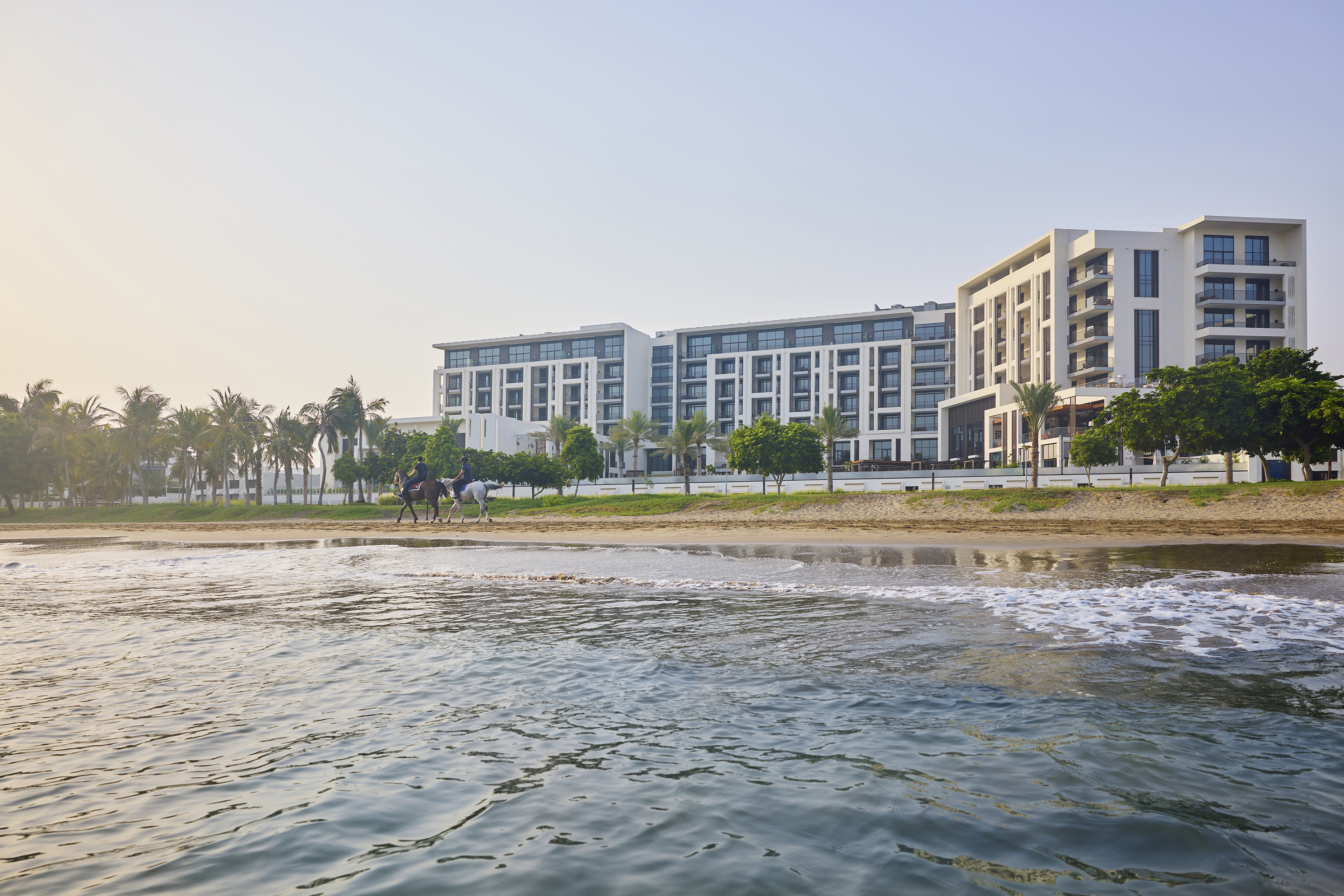 The sleek exterior of Mandarin Oriental Muscat with horse riders riding along the beach in front