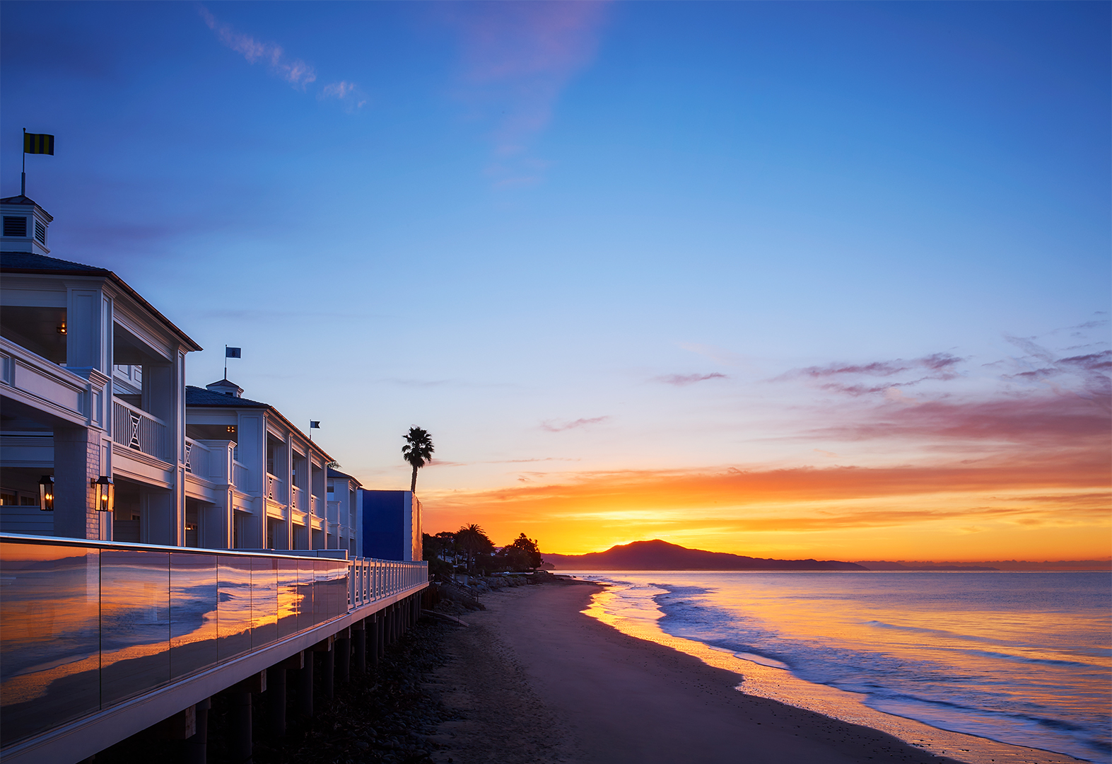 A serene sunset over a beach with an oceanfront building, palm trees, and distant mountains.